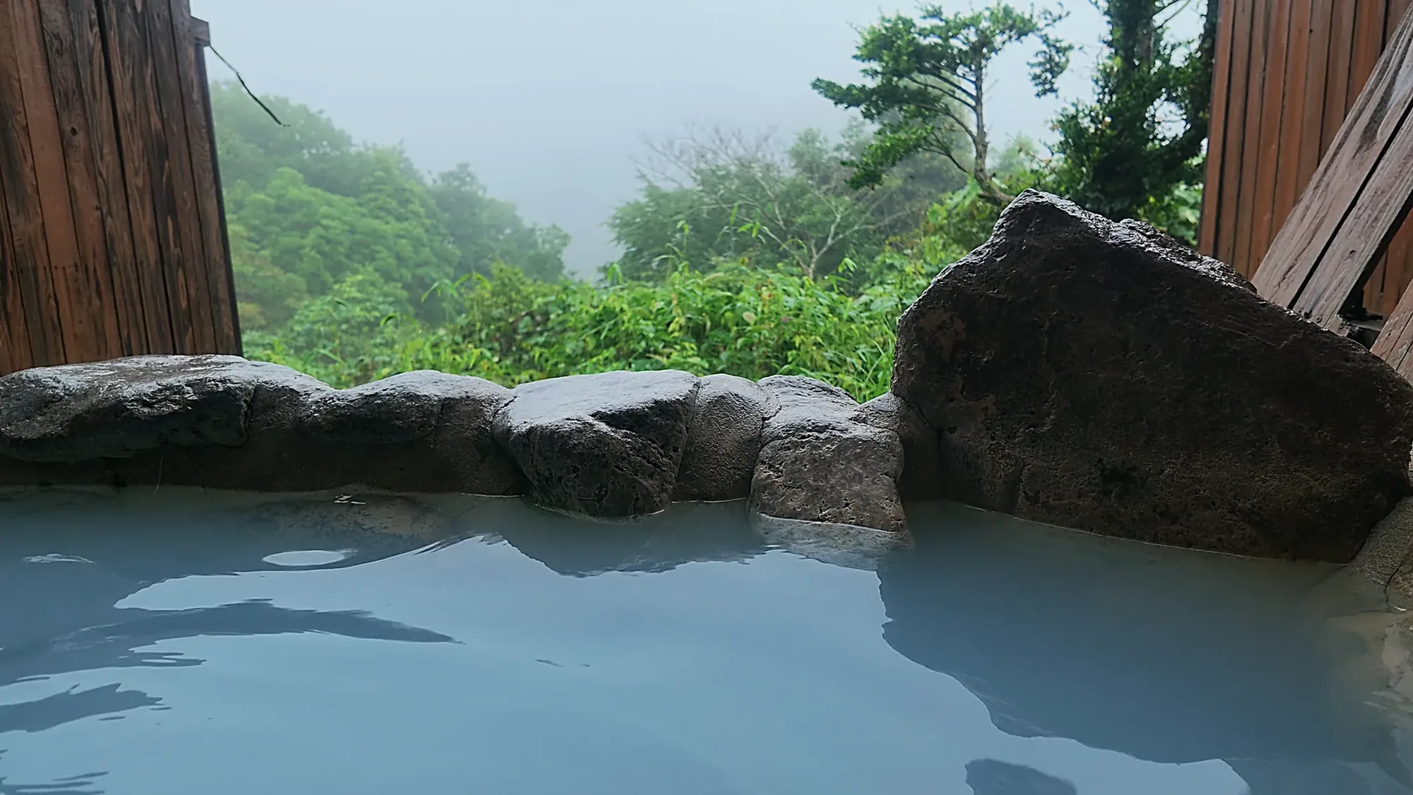 A milky hot-spring pool with a large boulder, enclosed by timber walls