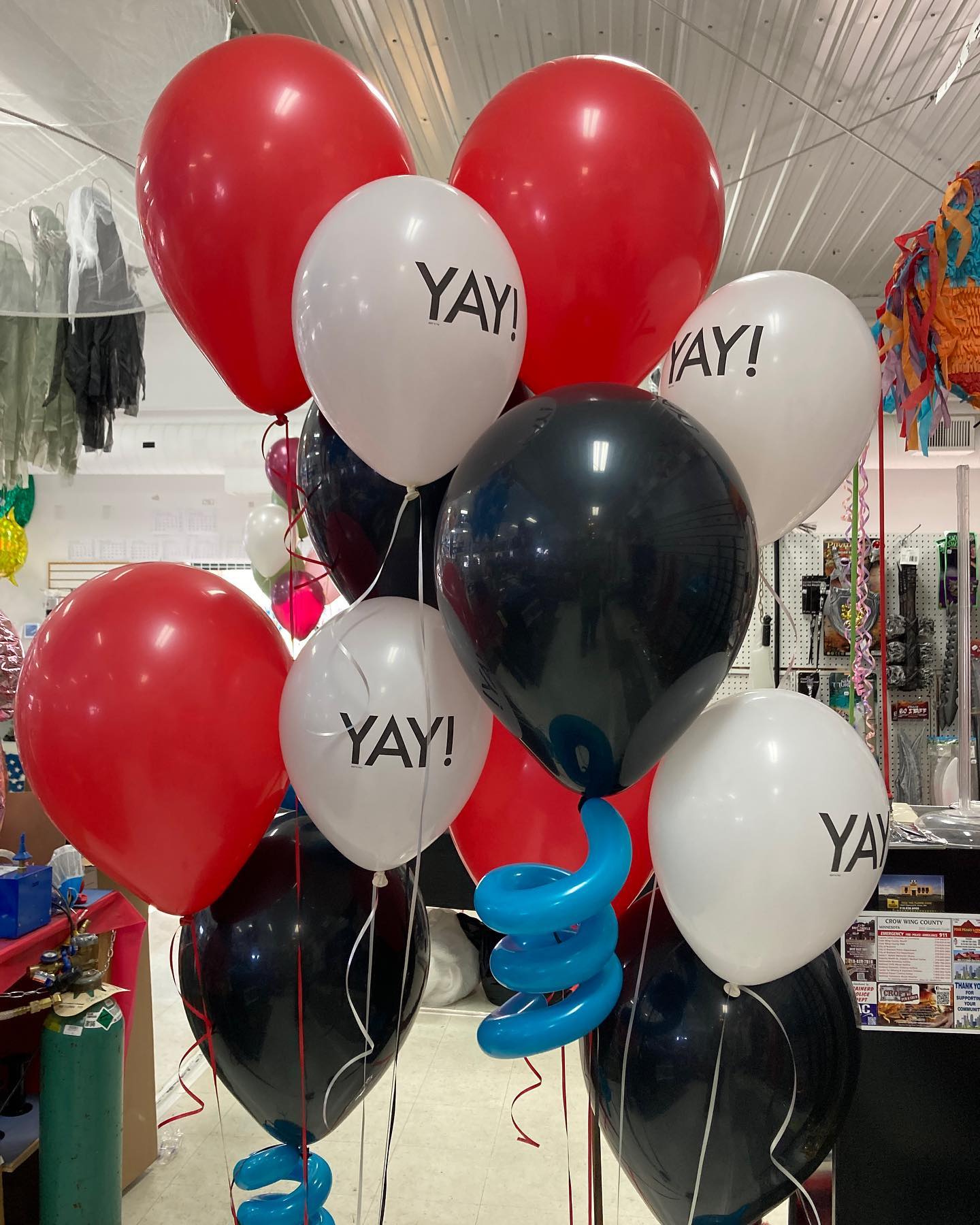A colorful cluster of helium balloons features red, black, and white balloons, some with "YAY!" printed on them, against a store backdrop with ceiling metal panels and various party supplies displayed on shelves.