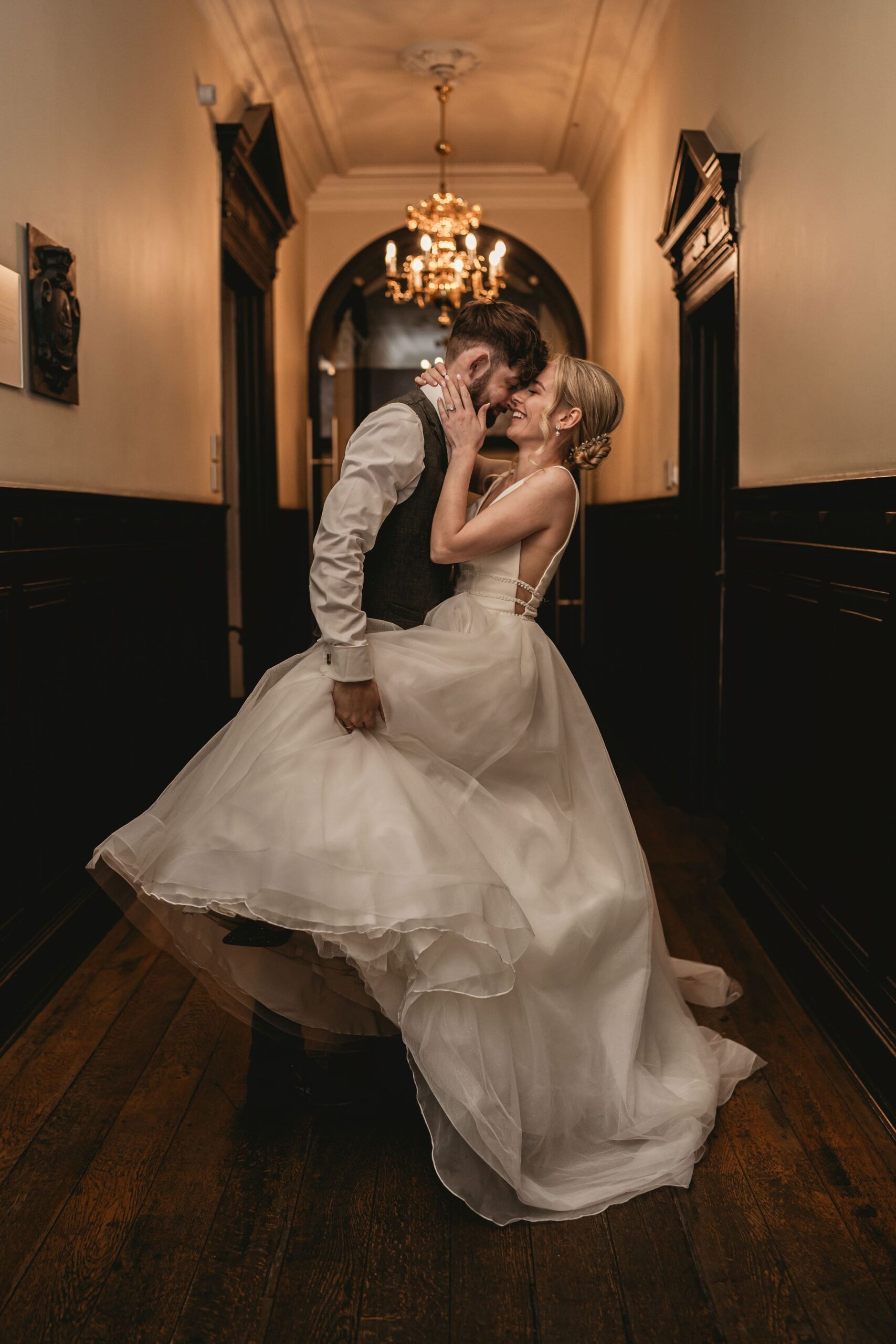 Brautpaar küsst sich in einem eleganten Flur vor einem Kronleuchter.