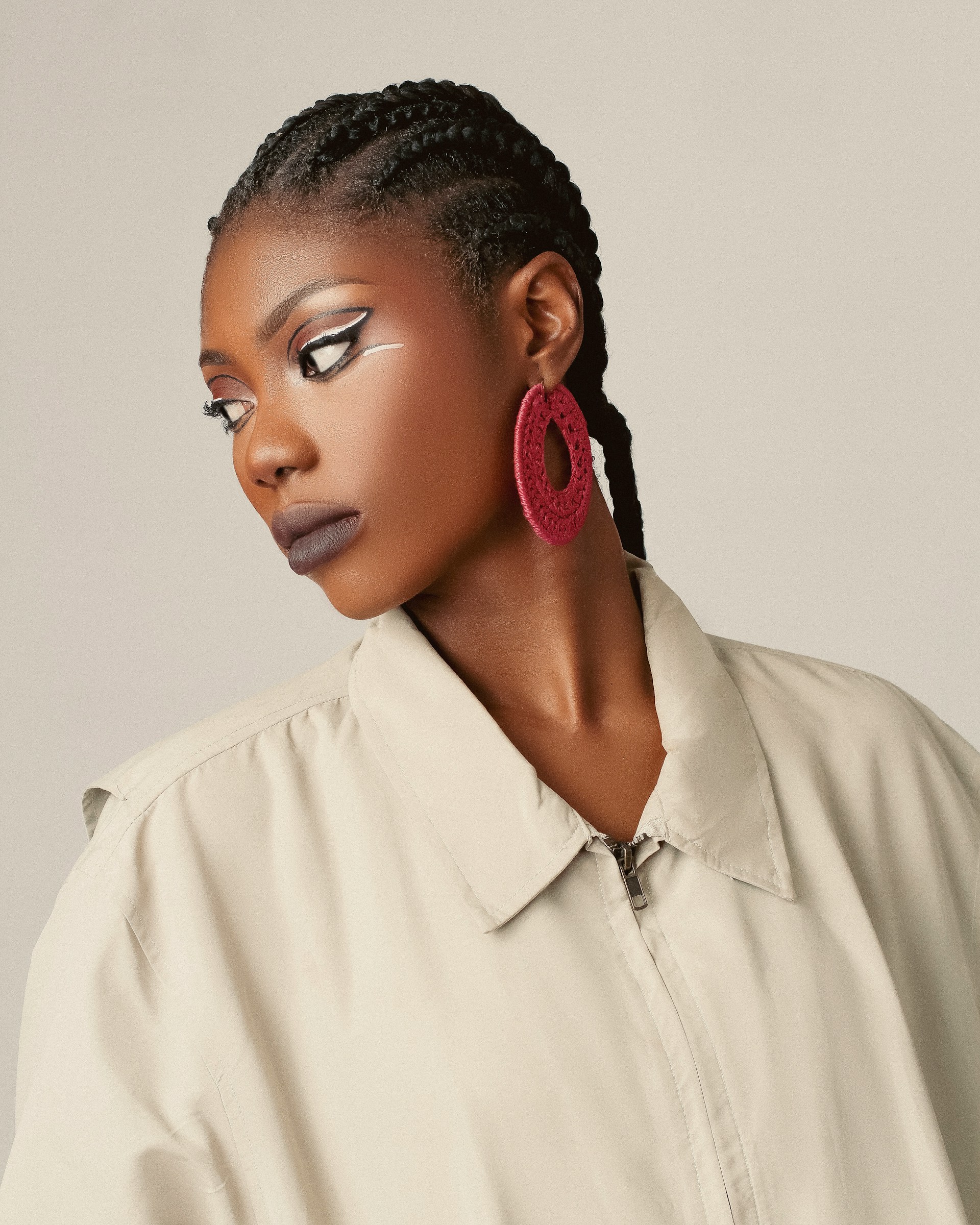 A Black woman with neat cornrow braids and bold graphic eyeliner looking over her shoulder, wearing a beige jacket and large pink circular earrings.