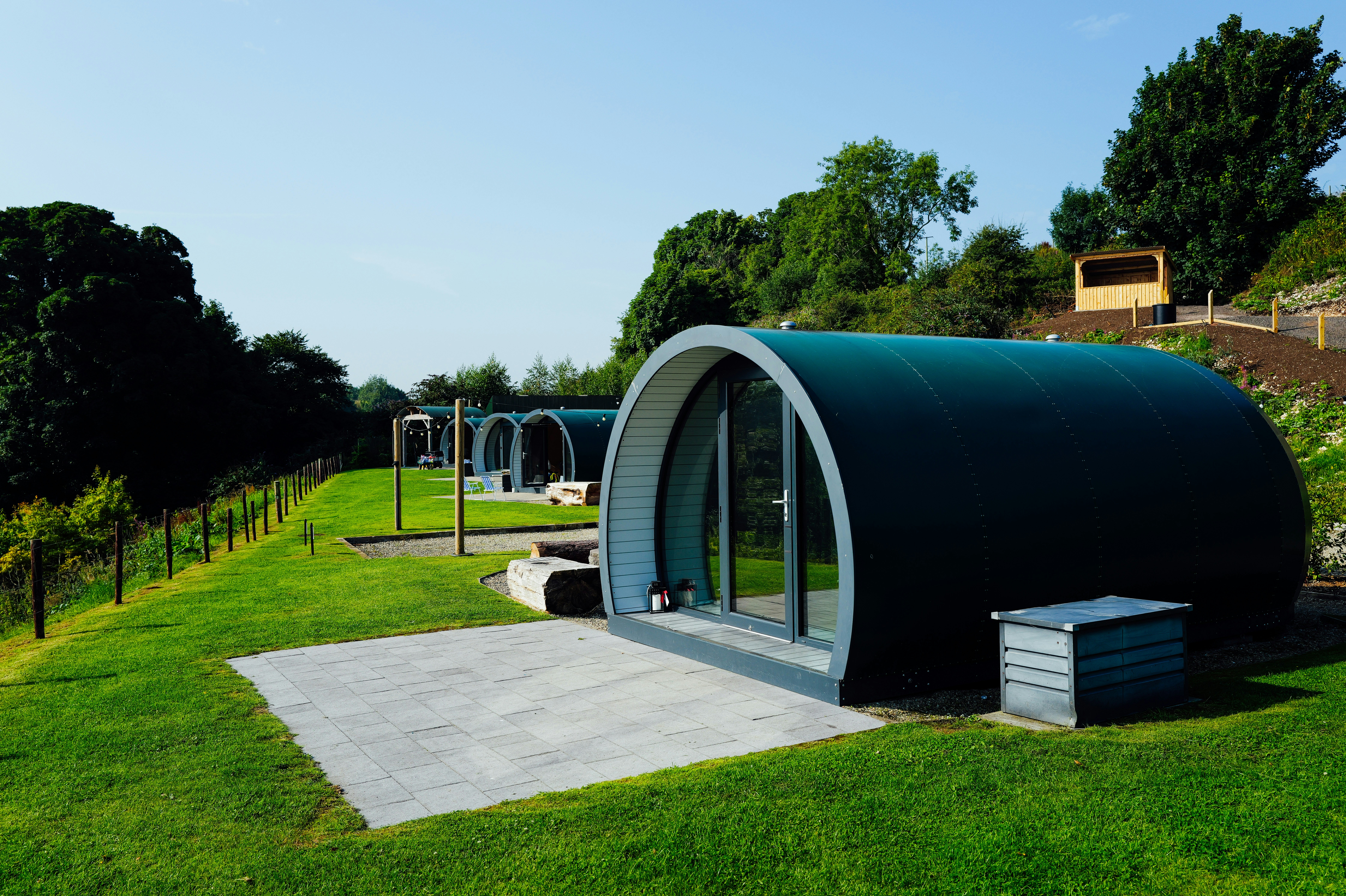 A row of modern, curved-roof glamping pods sits on a neatly mowed grassy hillside, with trees and a clear blue sky in the background.