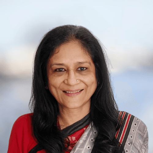 A smiling man with curly hair is sitting at a desk, looking at the camera, with a laptop and a lamp in the background.