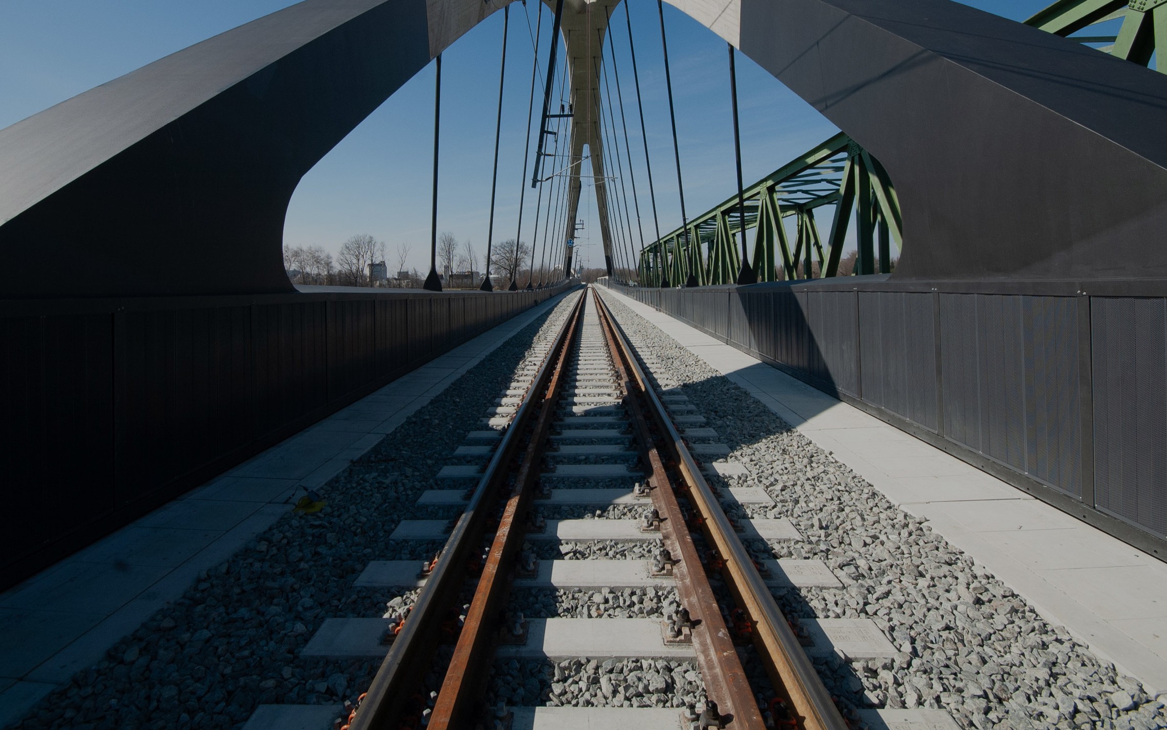 Gleise führen unter asymmetrischen, geschwungenen Stahlbögen einer modernen Brücke hindurch, klarer Himmel im Hintergrund.