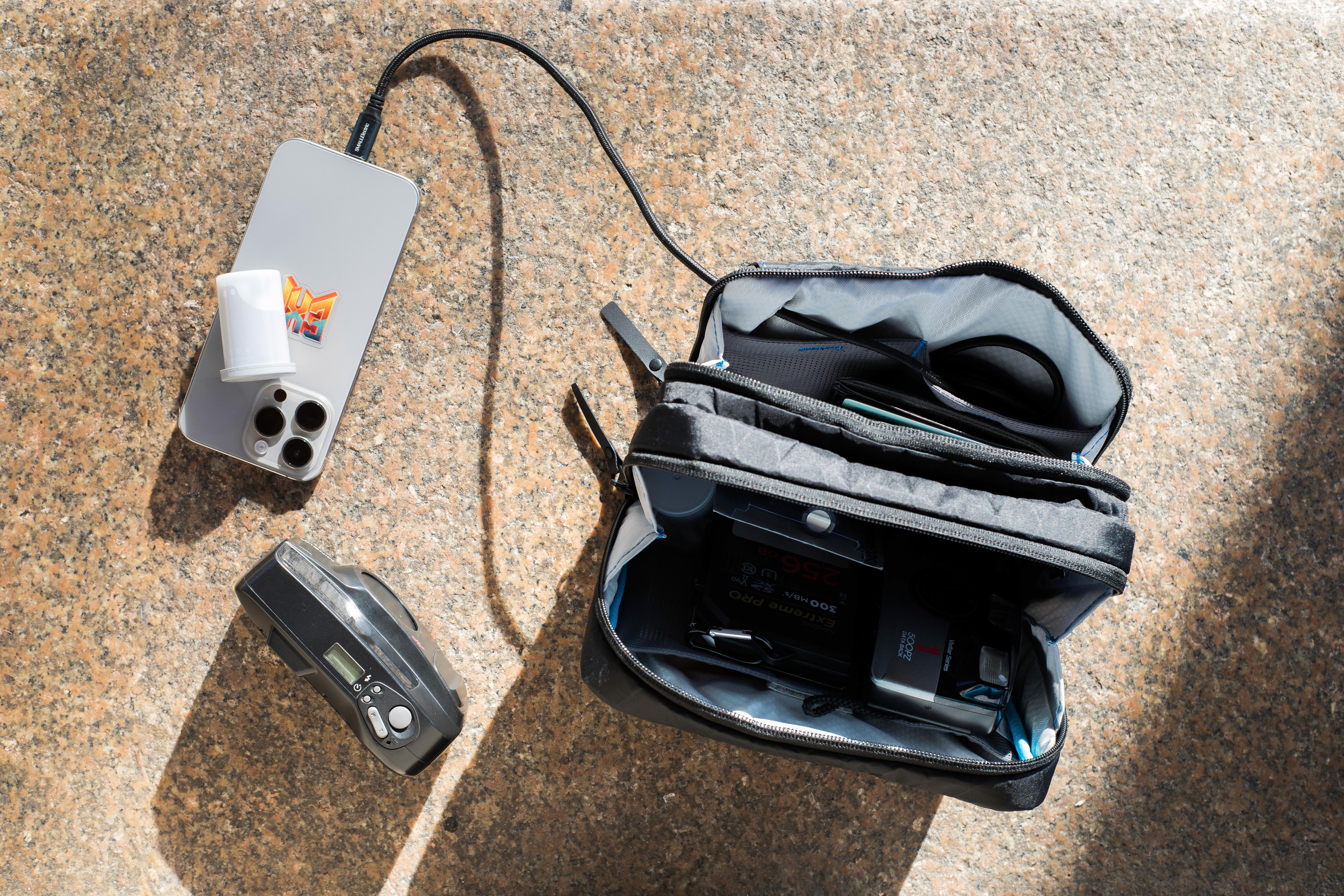 Open Gura Gear bag on pavement with an iPhone, compact camera, and accessories arranged around it.