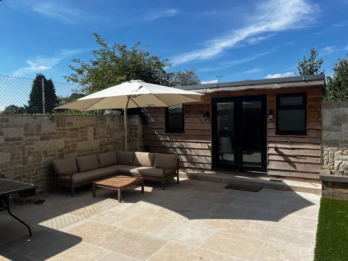 Paved garden patio with outdoor seating and a large parasol beside a timber garden room.