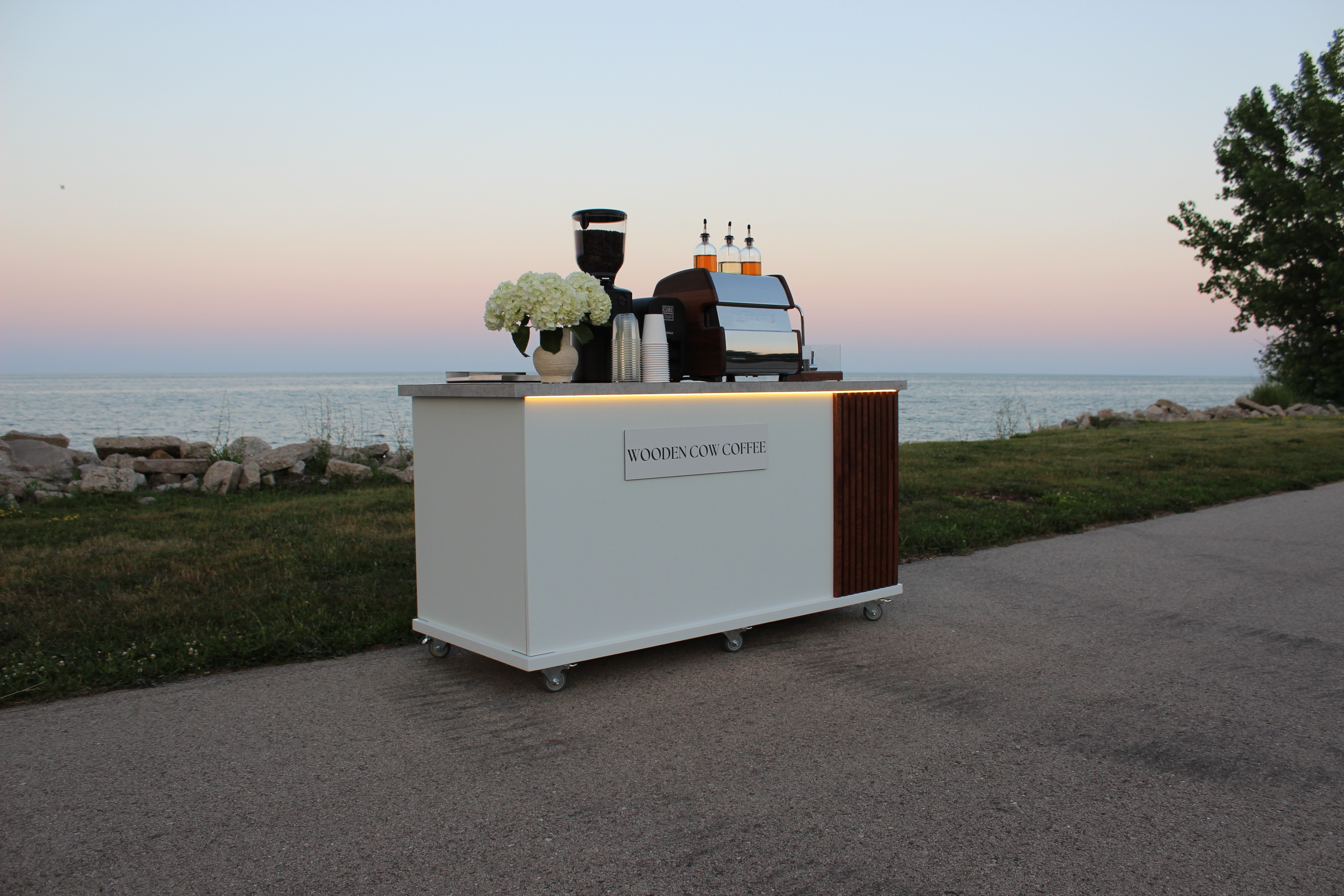 Black and white photo of a smiling man with glasses and a beard standing behind a mobile coffee setup outdoors. He is flanked by a coffee grinder filled with beans on the left, and an espresso machine with three syrup bottles on the right. A tamping device labeled “CUBE TAMP” is placed in the middle. Behind him, a grassy field with trees and a clear sky stretches into the distance, and string lights hang from a wooden post to the left.