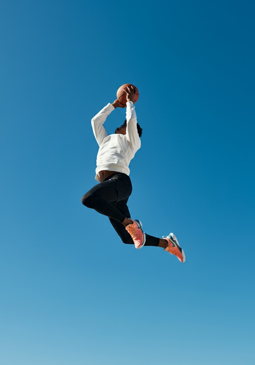 Athlete in mid-air with basketball, wearing white shirt and orange sneakers against a clear blue sky, showcasing dynamic motion.