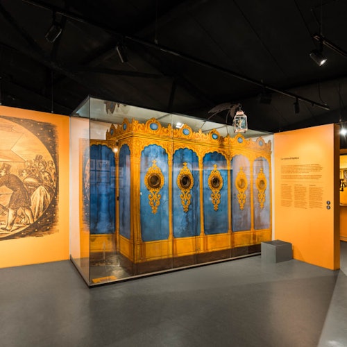 An ornate blue and gold carousel organ display in a museum with orange walls and spotlights illuminating the exhibit.