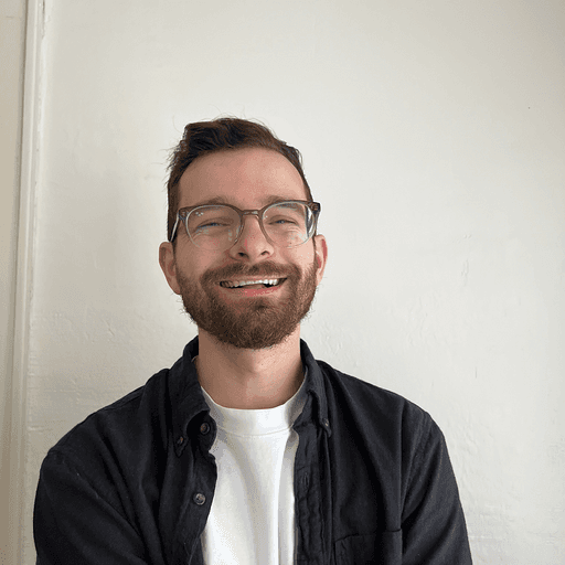 A man with dark hair and a beard is looking directly at the camera, wearing a black shirt against a simple background.