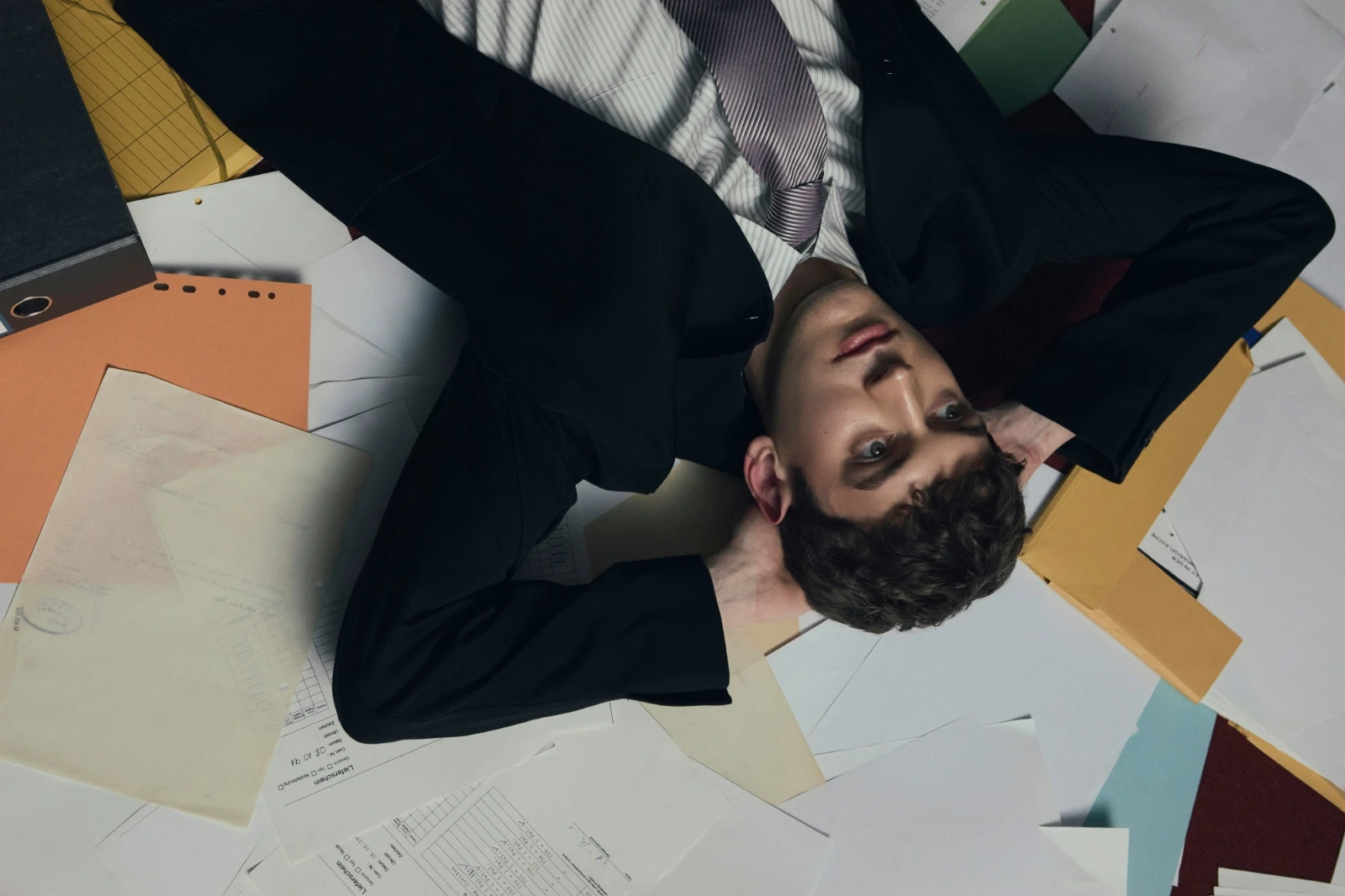 Ein erschöpft wirkender Geschäftsmann liegt zwischen ungeordneten Dokumenten und Akten auf dem Boden, den Blick nach oben gerichtet. Das Bild symbolisiert Informationsüberforderung, kognitive Last und den Wunsch nach automatisierter Klarheit und intelligenter Ordnung.