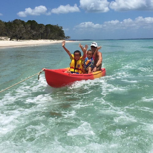 Three people in a small red boat on clear, shallow water near a sandy beach, waving and wearing life jackets.