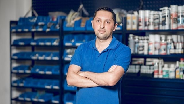 Homem de camisa azul de braços cruzados; ao fundo, prateleiras organizadas com gaveteiros azuis e latas de produtos.