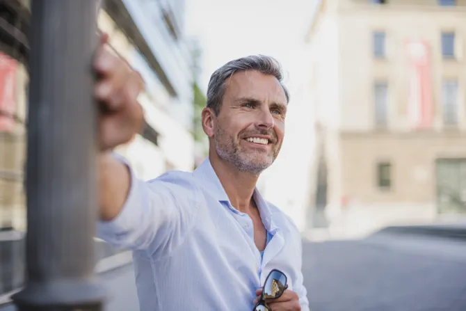 Smiling middle-aged man in a light blue shirt leaning against a lamp post in a sunny urban setting.