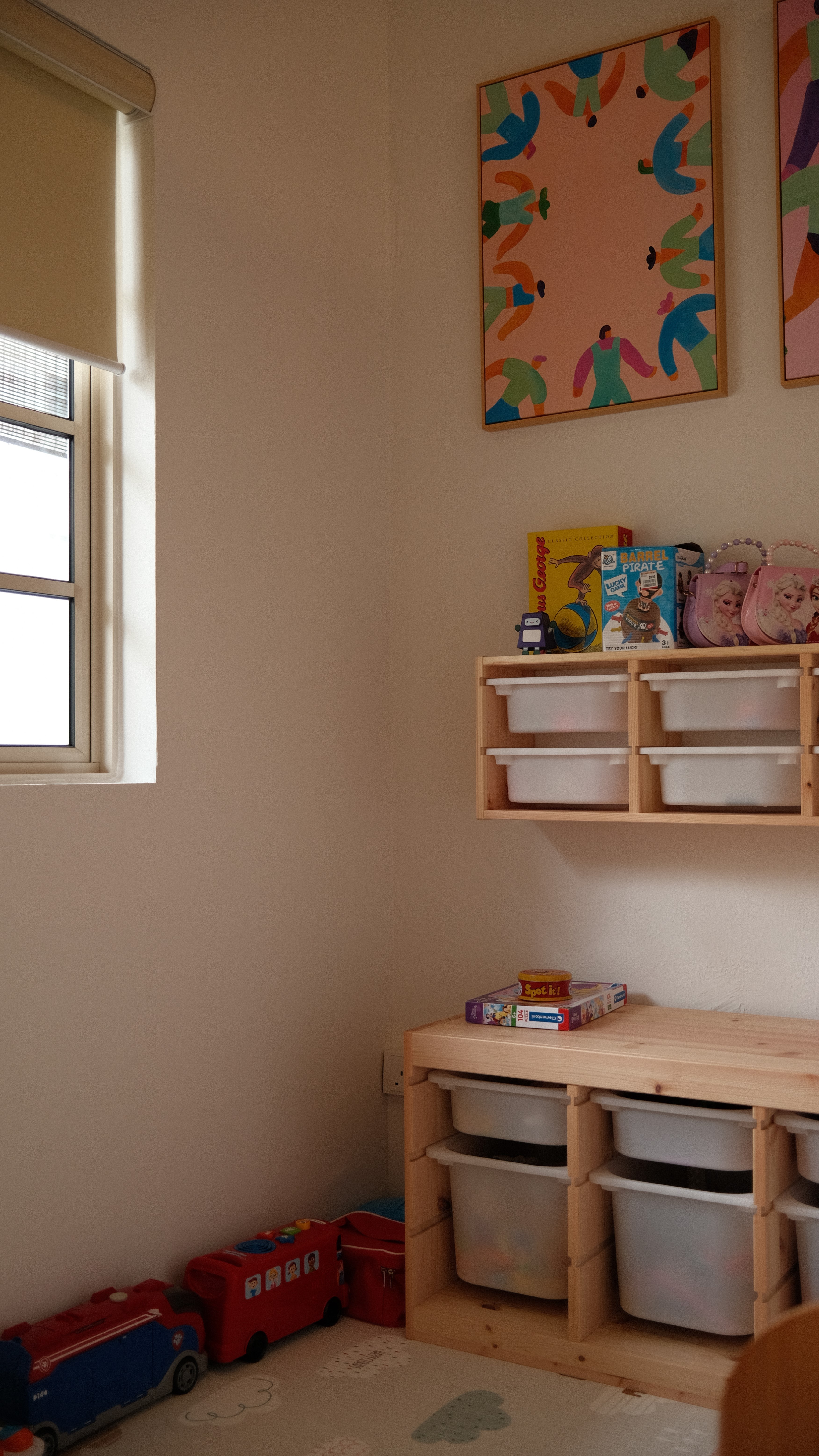 Children’s playroom corner with natural light, pastel-colored abstract art on the wall, and organized wooden toy storage bins beneath a window.