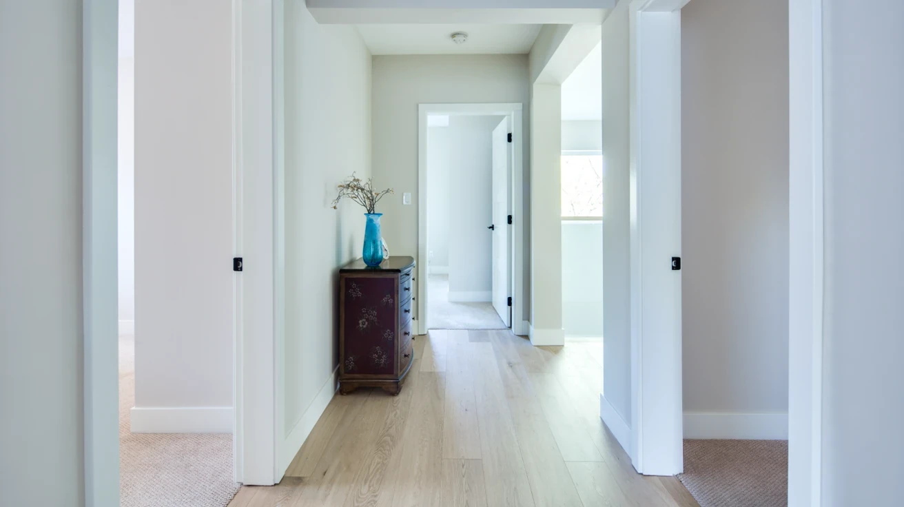 Upstairs hallway with light hardwood flooring, soft blue-grey accent walls, white trim, and access to multiple bedrooms in custom built home