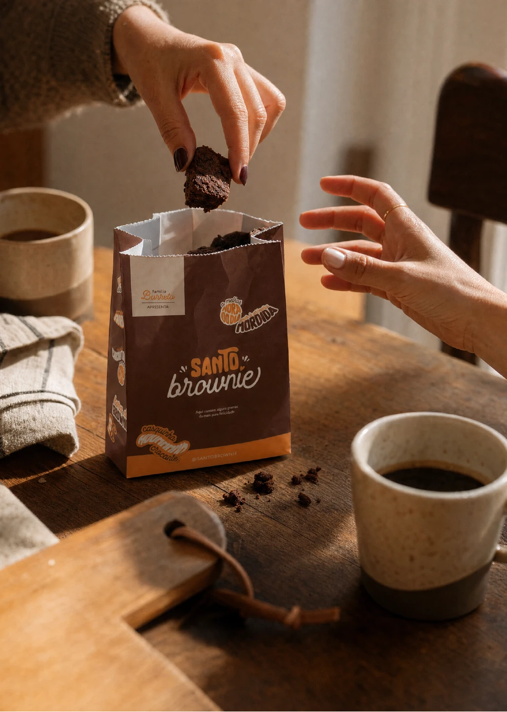 Two hands reaching into a brown kraft paper bag printed with the Santo Brownie logo, set on a wooden table beside two cups of coffee.