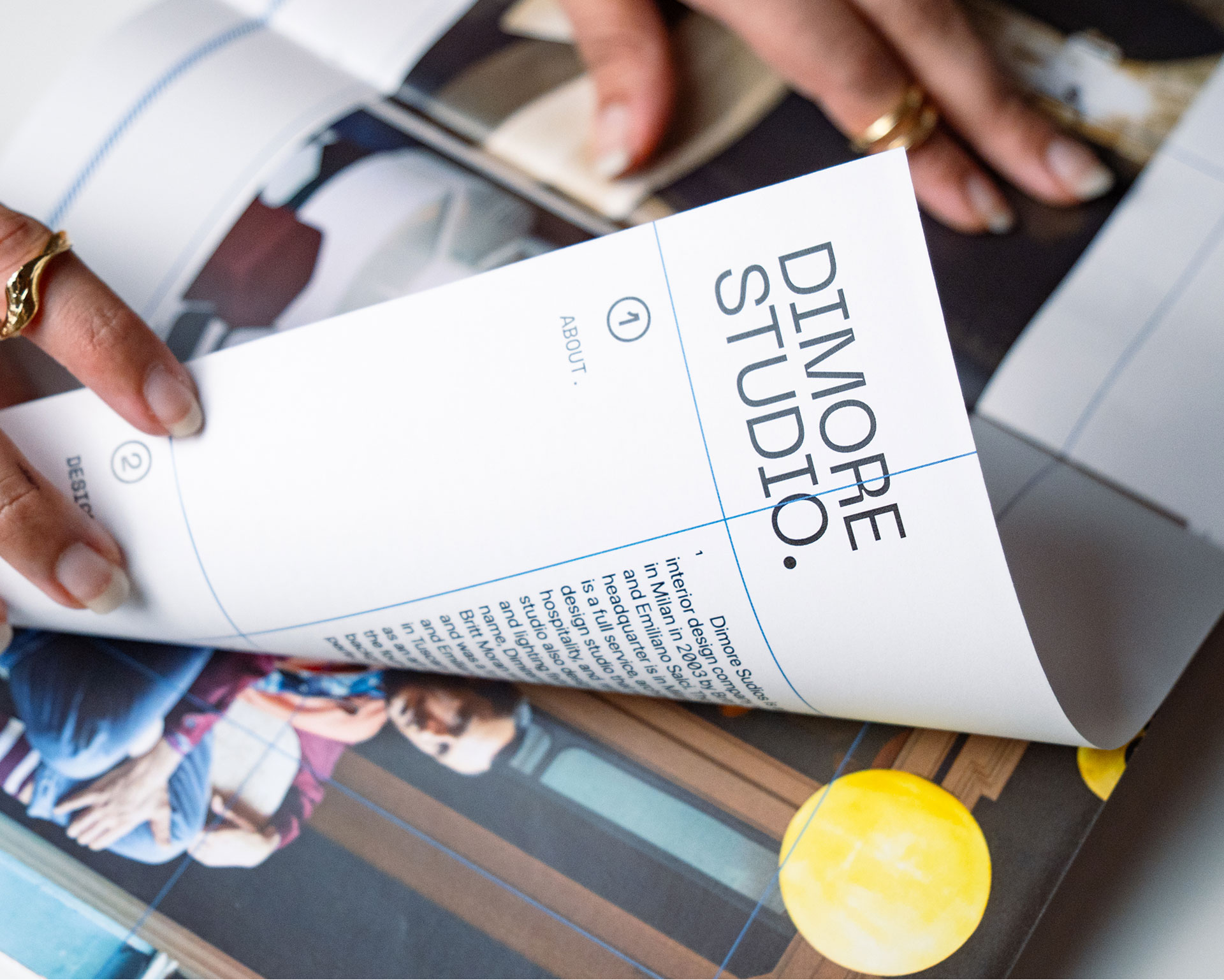 Hands flipping through a glossy magazine displaying a spread with graphics and articles, featuring a page titled "DIMORE STUDIO" with text and a modern design layout.