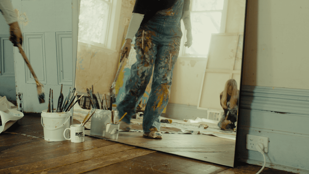 A painter standing and holding a brush, with vibrant paintings covering the walls and floor, adding the final touches to their work