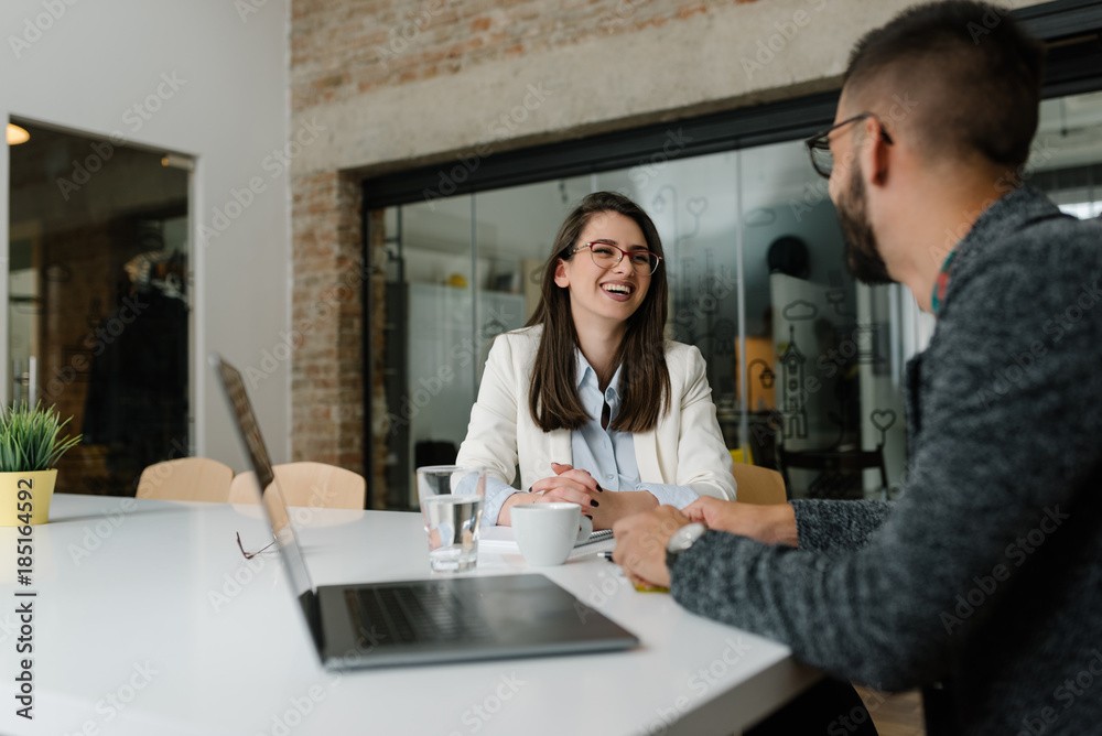 Two professionals smiling and engaging in a conversation during a meeting, with a laptop and a glass of water on the table.