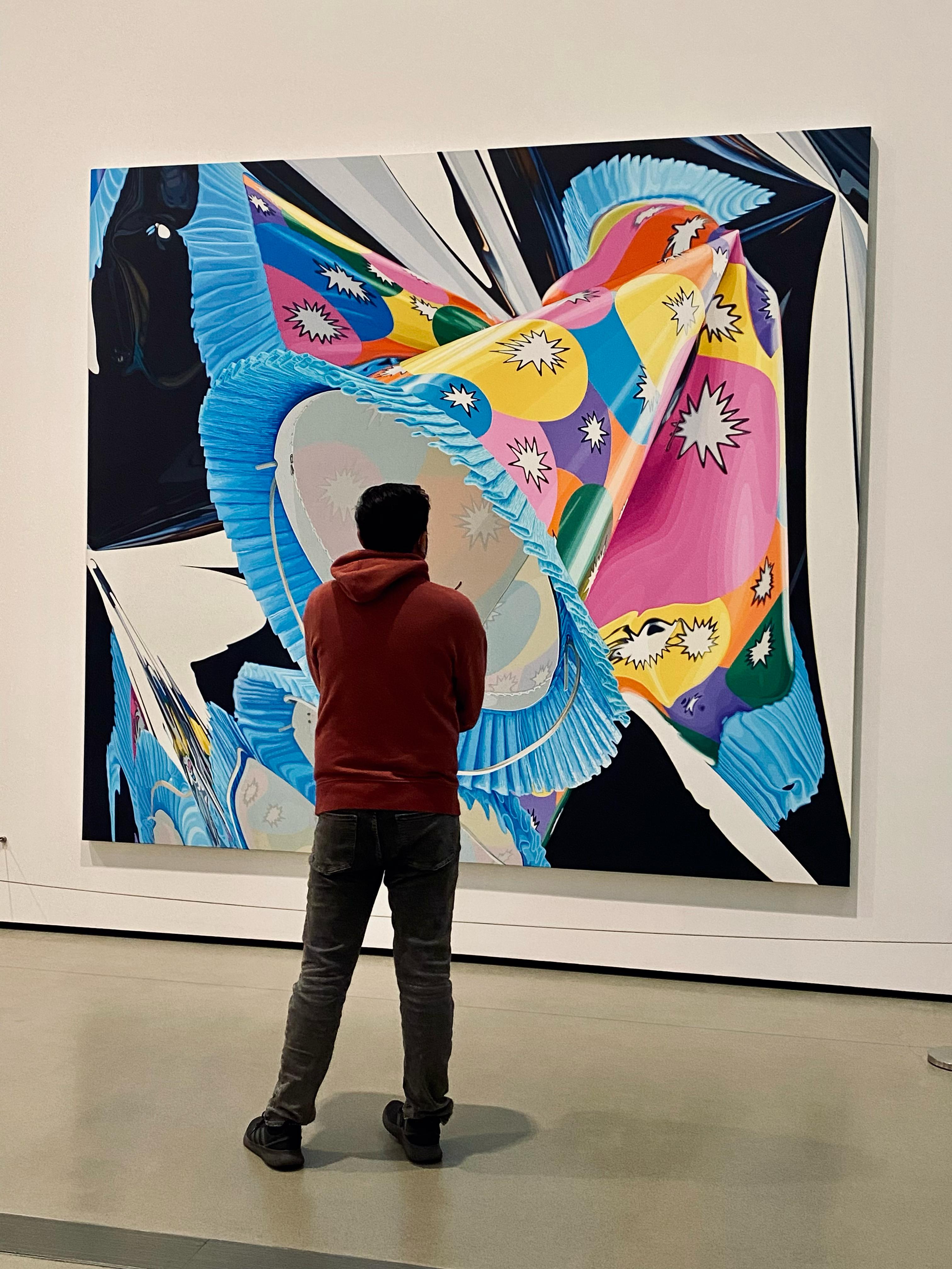 Amish in a red hoodie stands in front of a large, colorful abstract painting in an art gallery.