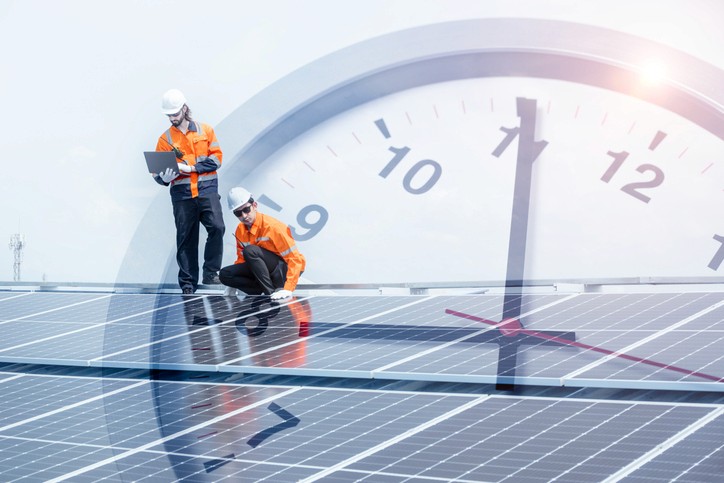 Zwei Arbeiter inspizieren Solarpaneele unter einem hellen Himmel, mit einer eingeblendeten Uhr als Symbol für Zeit und Effizienz.
