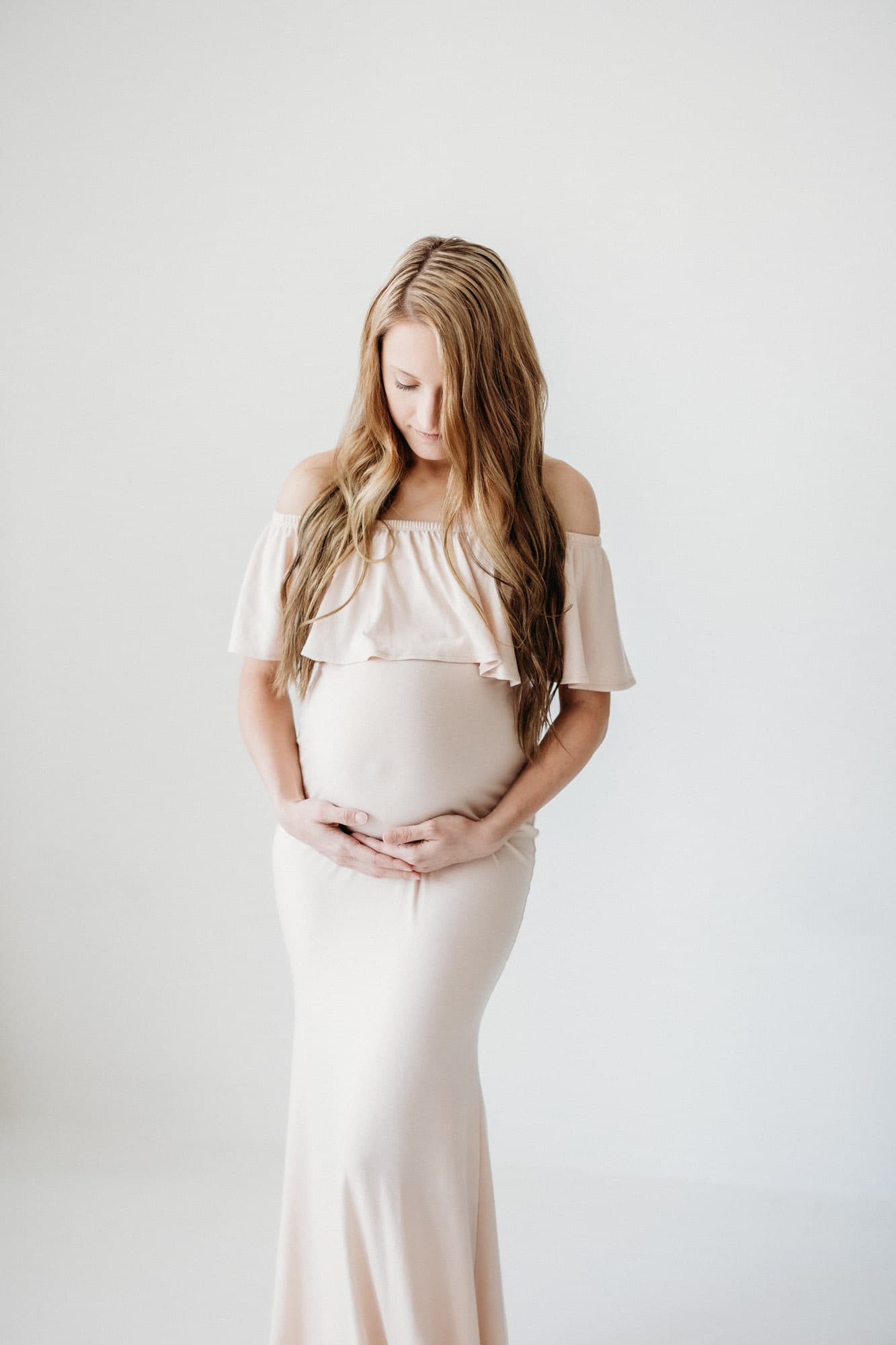 A mother-to-be in a floral dress holds her pregnant belly in the soft natural light of Revelator Studio in Shreveport, showcasing maternity photography.