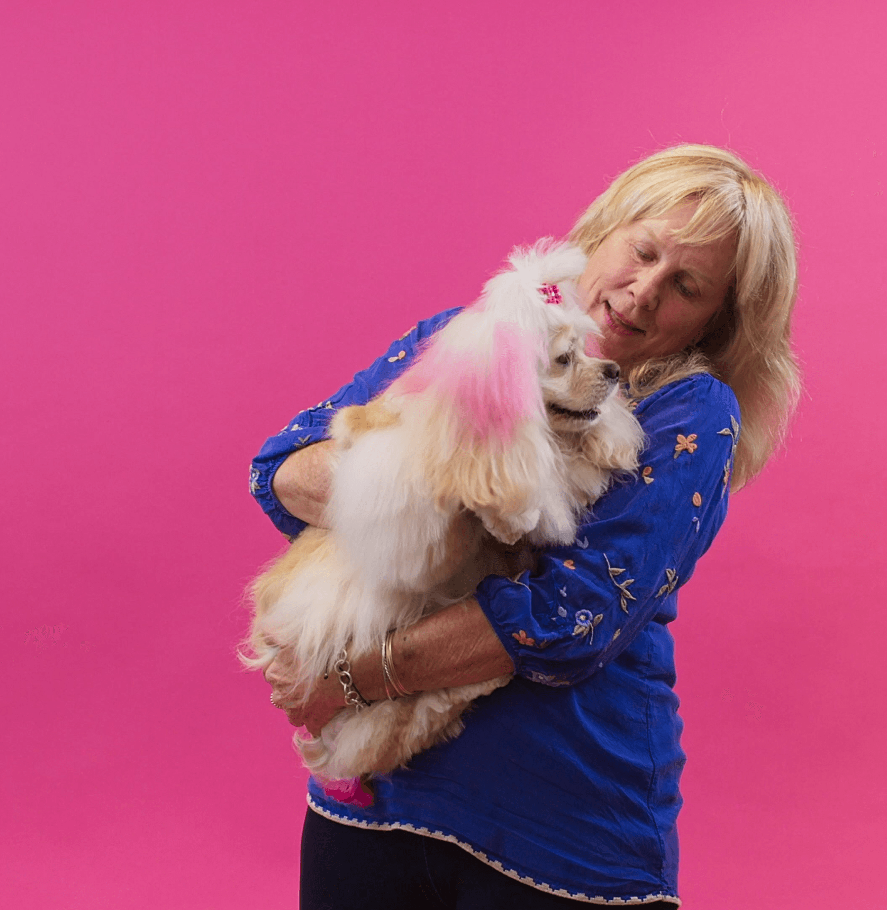 Smiling woman holding her fluffy white dog against a pink background to express love, care, and joyful grooming experience.