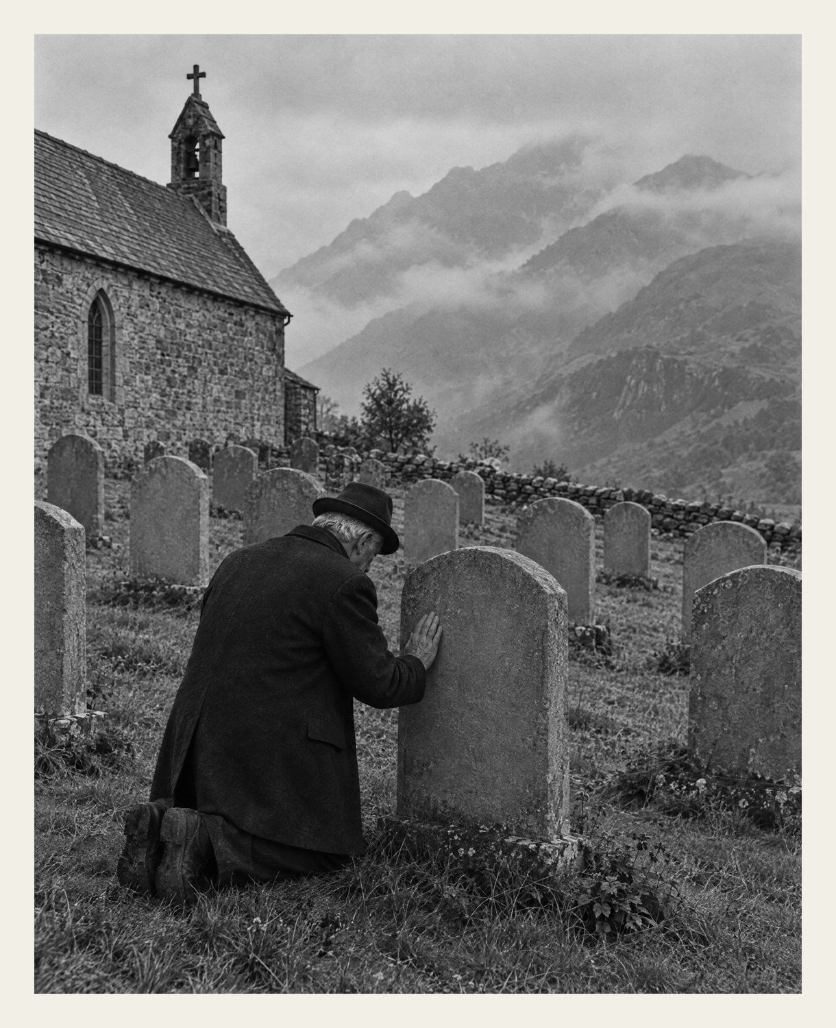 Black-and-white print of an old man kneeling at a blank gravestone in a mountain churchyard