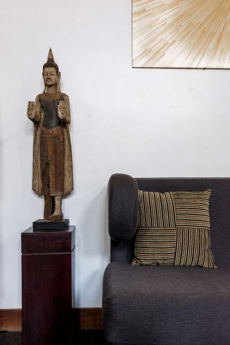 A tall wooden Buddha statue stands on a dark pedestal next to a gray sofa with a striped yellow and black pillow.