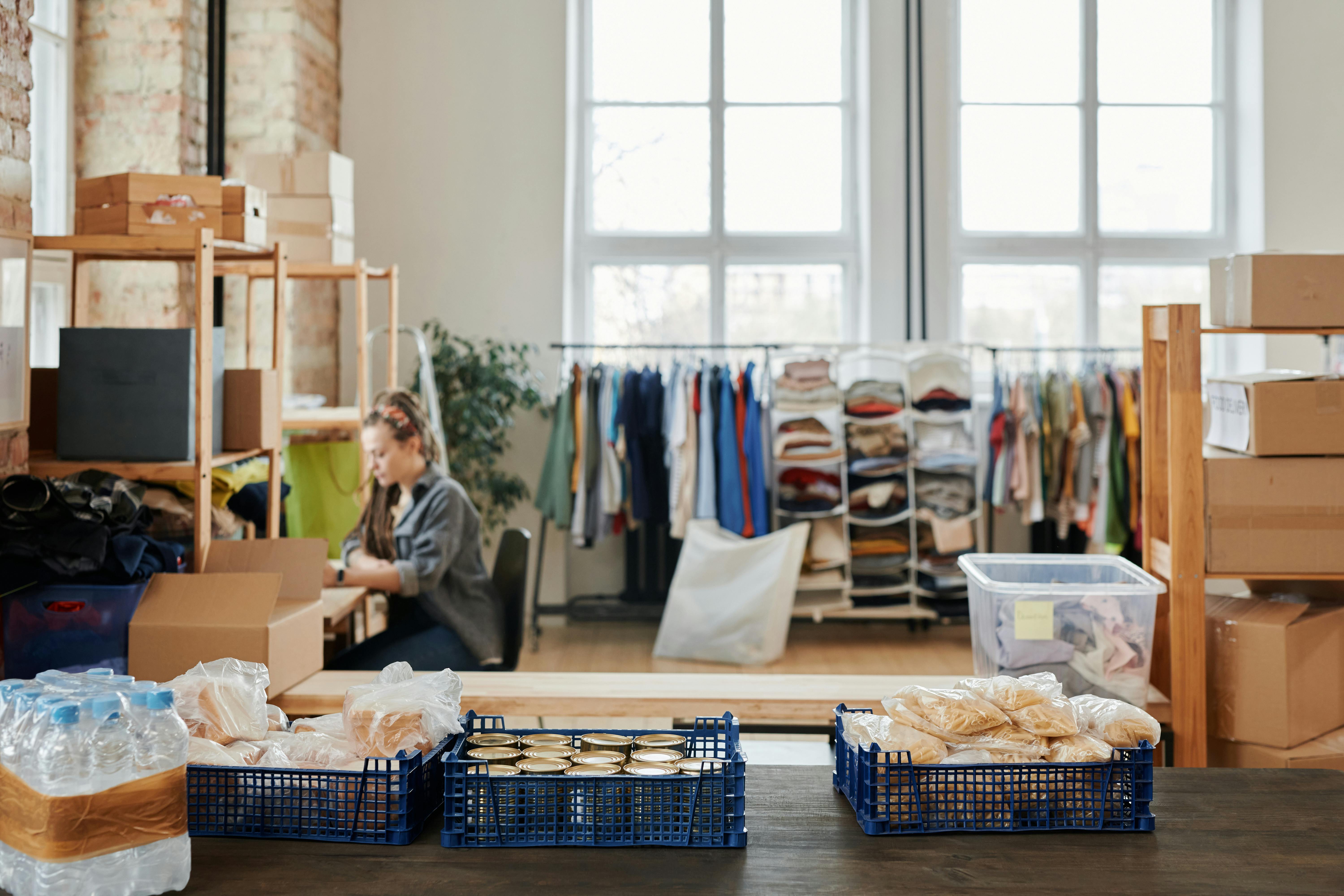 Vrouw sorteert hulpgoederen (voedsel, kleding) in een opslagruimte met grote ramen, dozen en houten rekken.