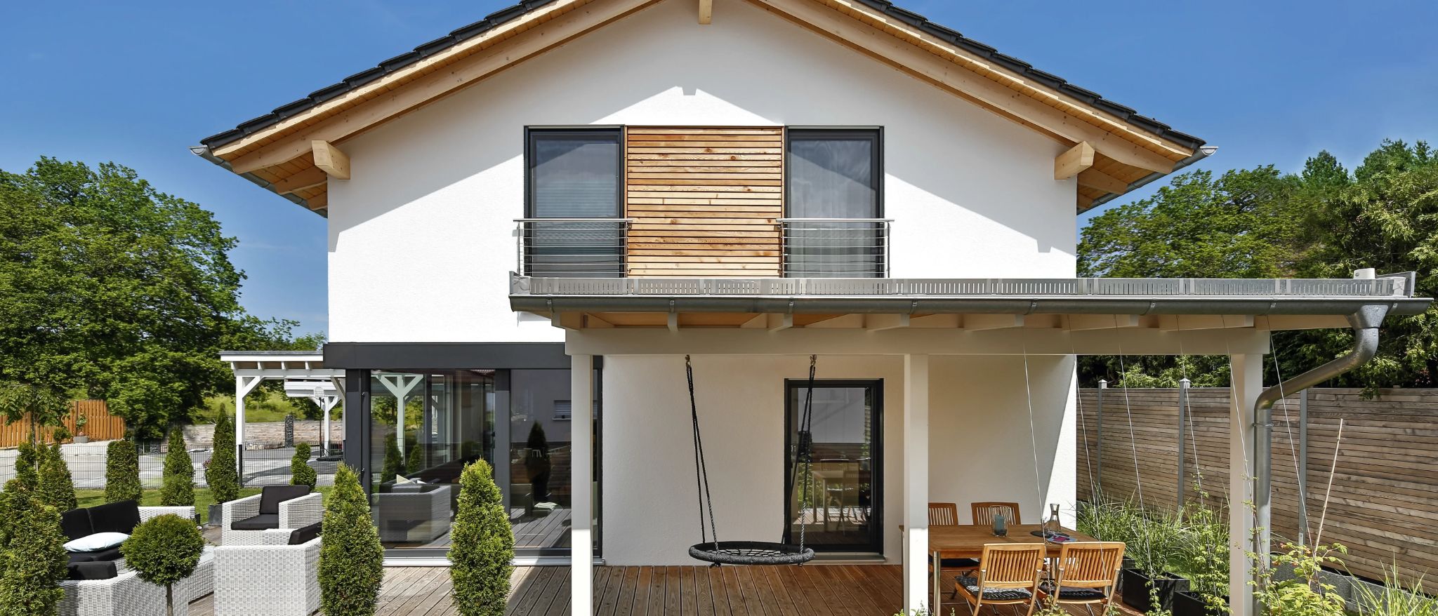 Moderne weiße Hausfassade mit Holzakzenten, Balkon mit Edelstahlgeländer und Sitzgruppe auf Holzterrasse.