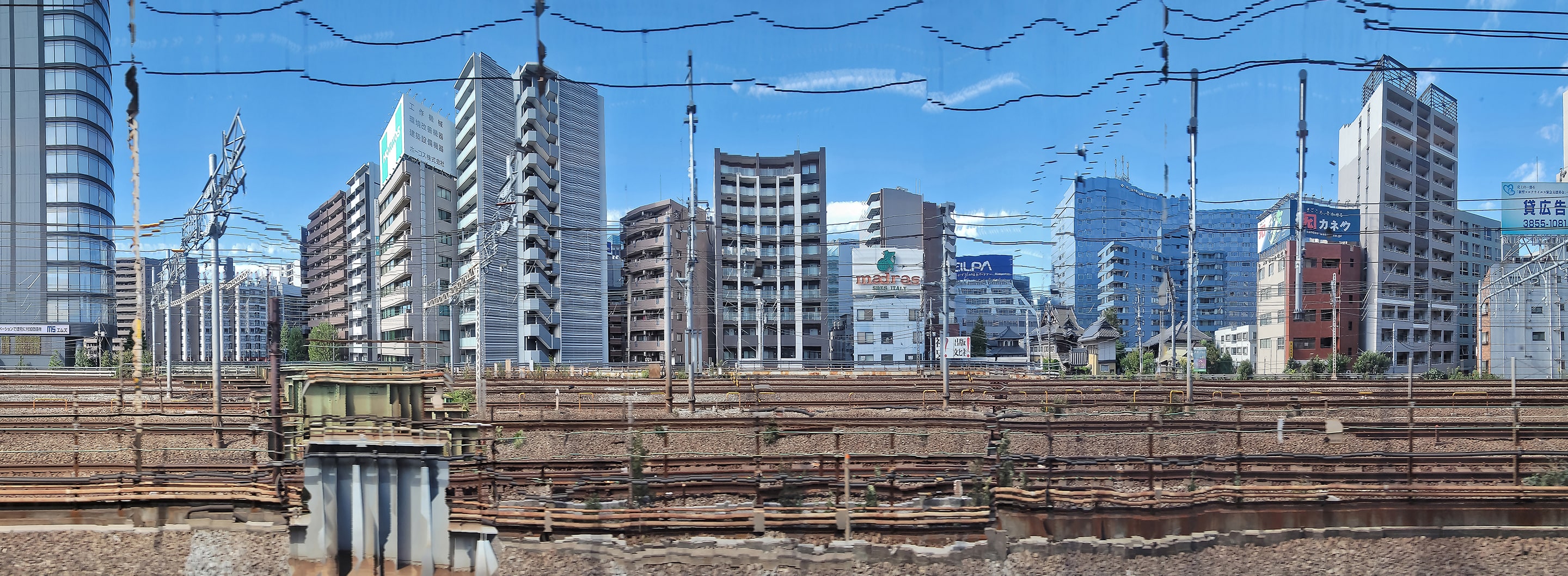Panoramic photograph of urban train tracks lined with high-rise buildings under a clear blue sky. The vertical lines of cables and architecture appear subtly warped, merging industrial structure with rhythmic visual distortion.