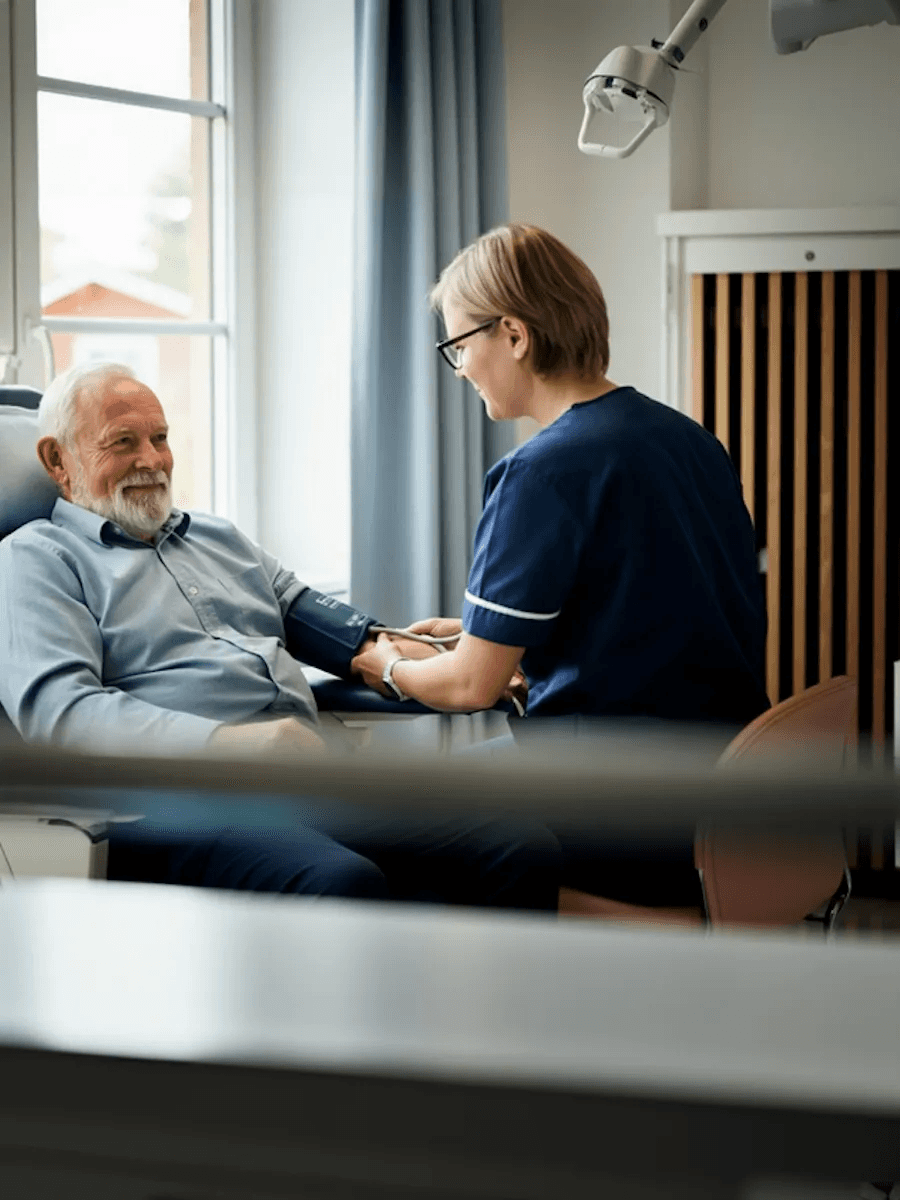 Nurse in navy scrubs taking an older man’s blood pressure in a medical consultation room.