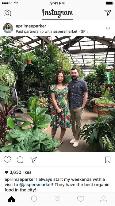 An Instagram post by 'aprilmaeparker' showing a woman in a floral dress and a man in a casual shirt standing among lush green plants in a garden center, tagged as a "Paid partnership with jaspersmarket." The caption promotes Jasper's Market for organic food, indicating influencer marketing.