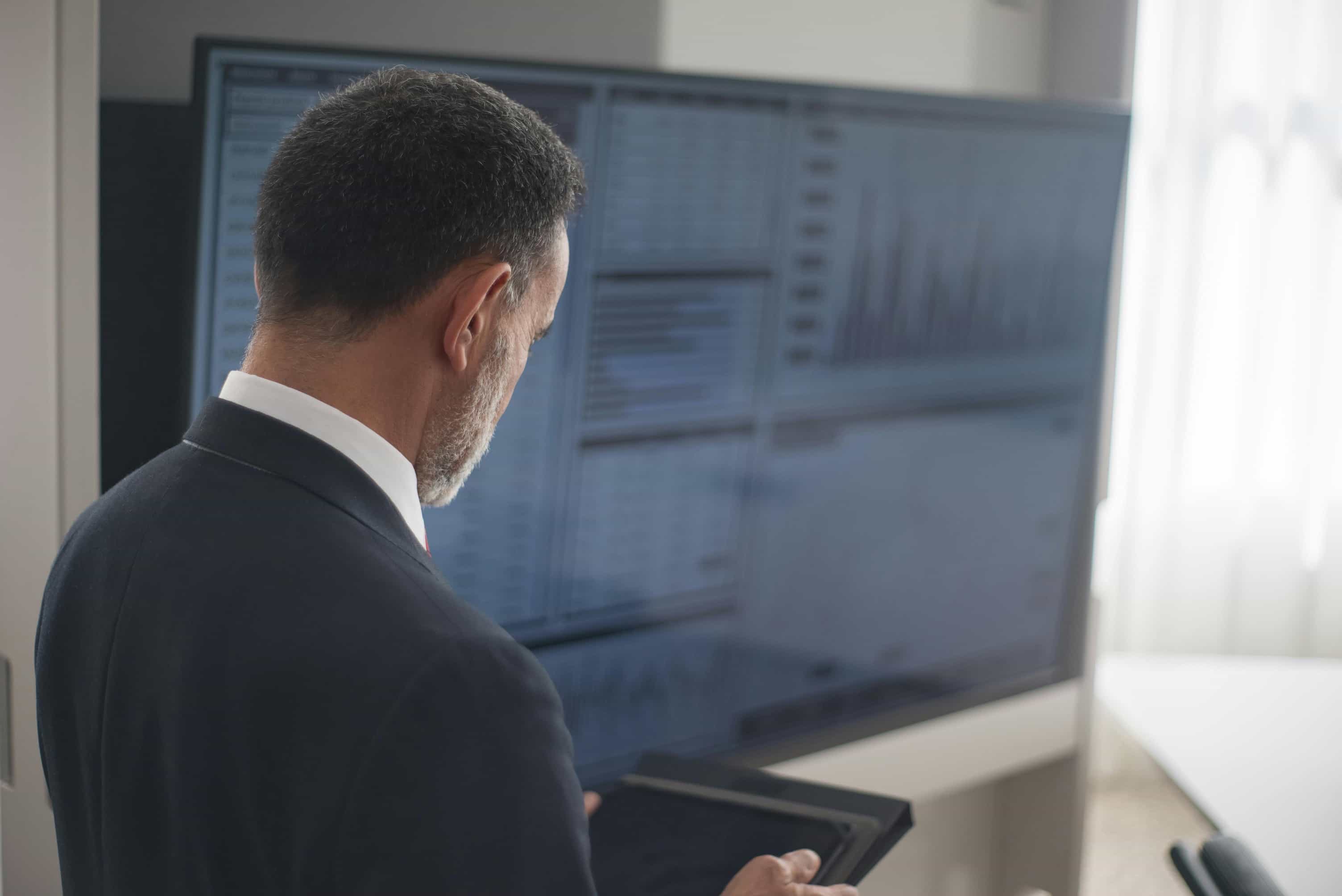 A man in a dark suit is seen from behind, looking intently at a computer monitor displaying graphs and charts in a bright office environment. He appears to be holding a mobile device or a tablet in his left hand.