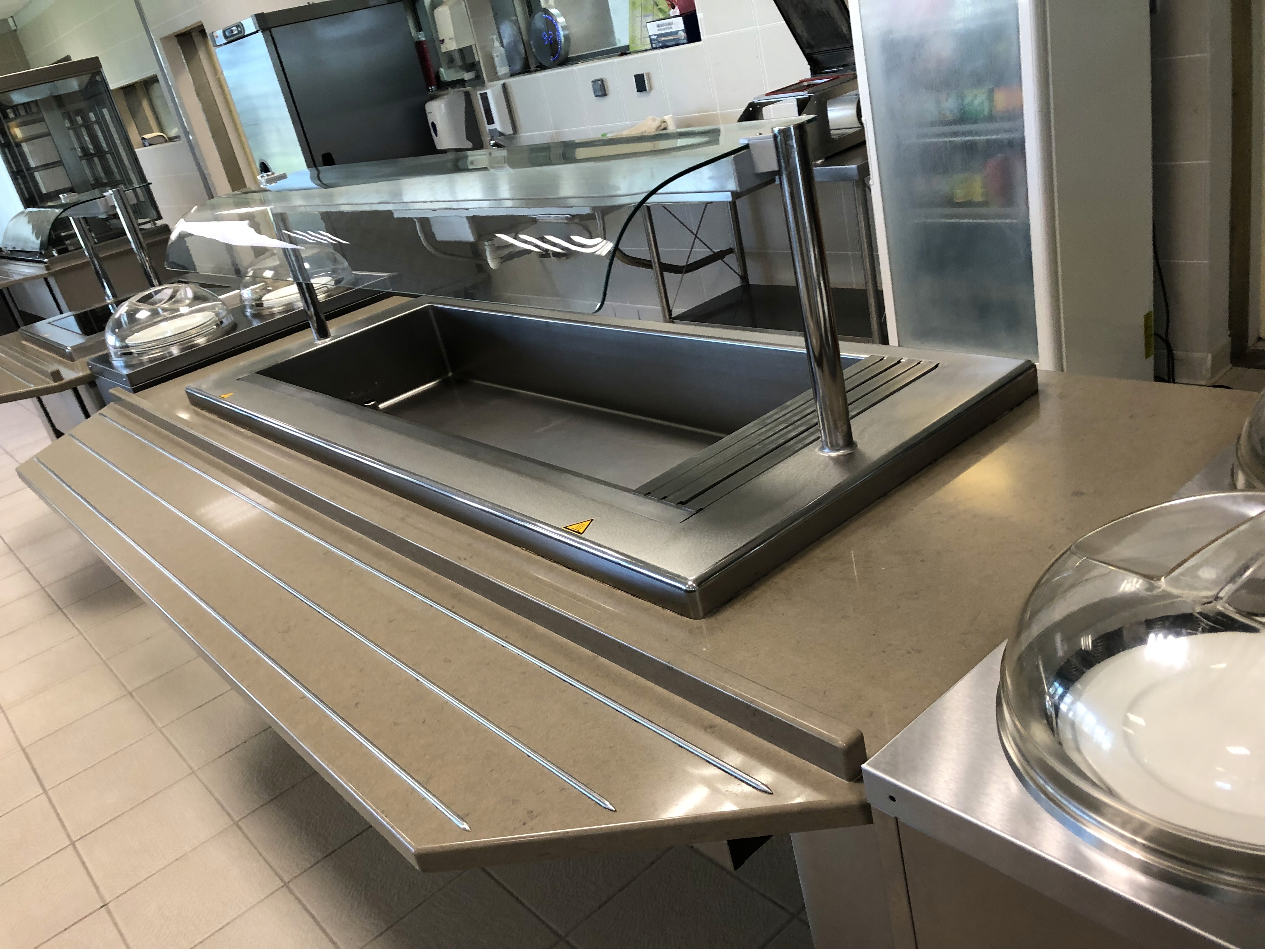 Comptoir de restauration en acier inoxydable, sans étagères, équipé d'une vitre de protection et de plateaux de service, dans un cadre de cafétéria moderne.