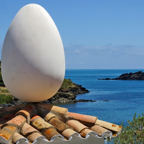 A large white egg sculpture sits atop a tiled roof, overlooking a coastal landscape with calm blue water and rocky shores.