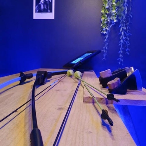 Close-up de uma mesa de madeira com dois arcos de tiro com arco e um tablet. A parede ao fundo tem plantas penduradas e uma foto emoldurada.