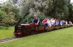 Families enjoying miniature railway ride through lush green parkland on open-air tourist train with scenic surroundings