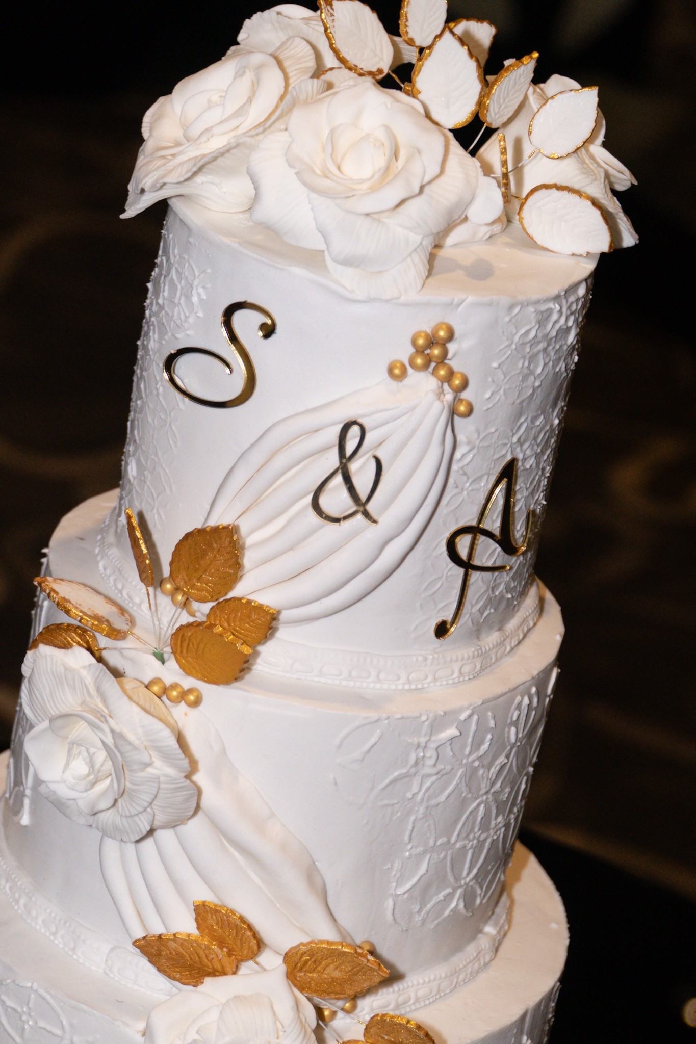 Two-tier white wedding cake decorated with white roses, gold leaves and gold lettering.