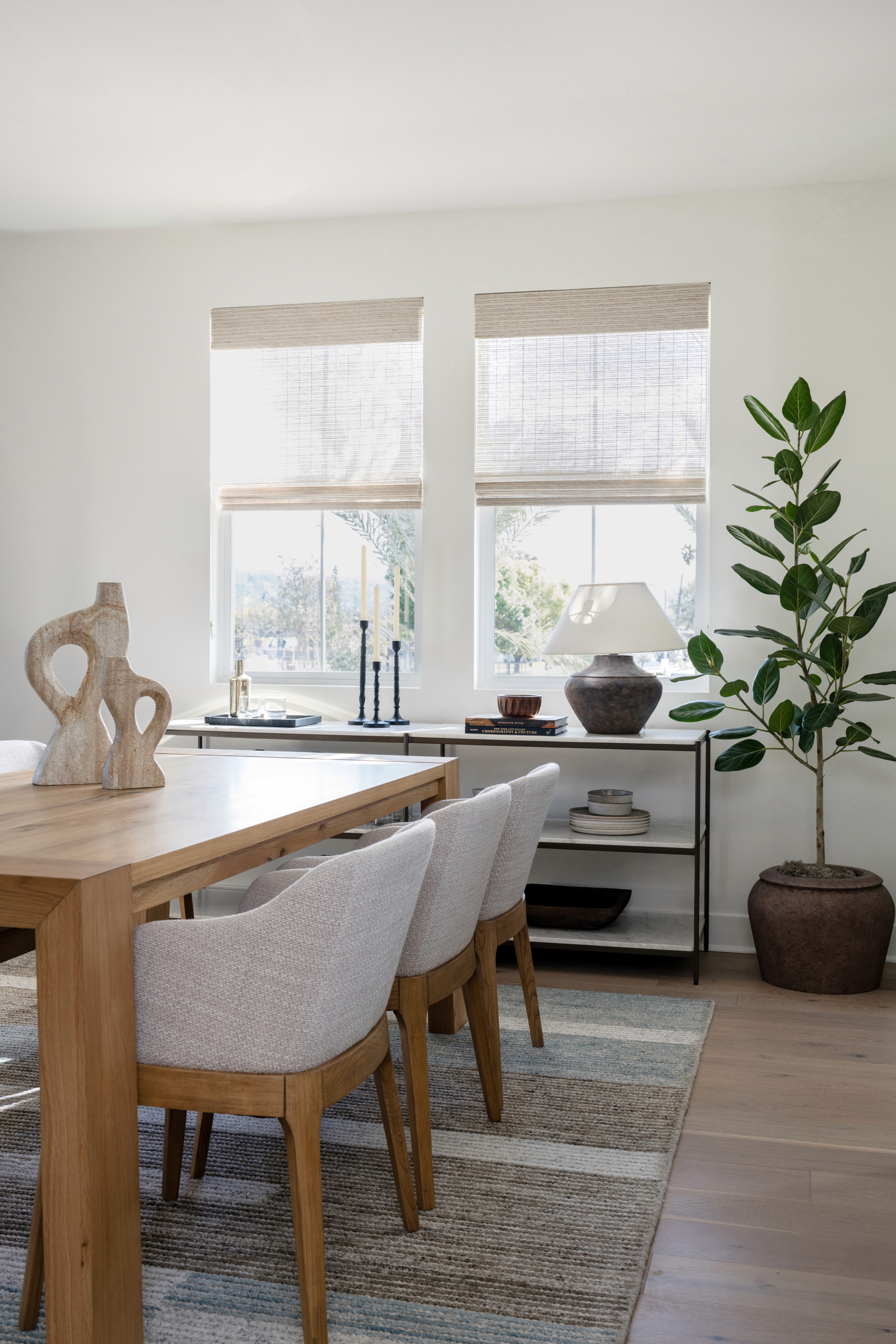 Light wood dining table table with upholstered chairs, open shelf wet bar under window and large potted plant