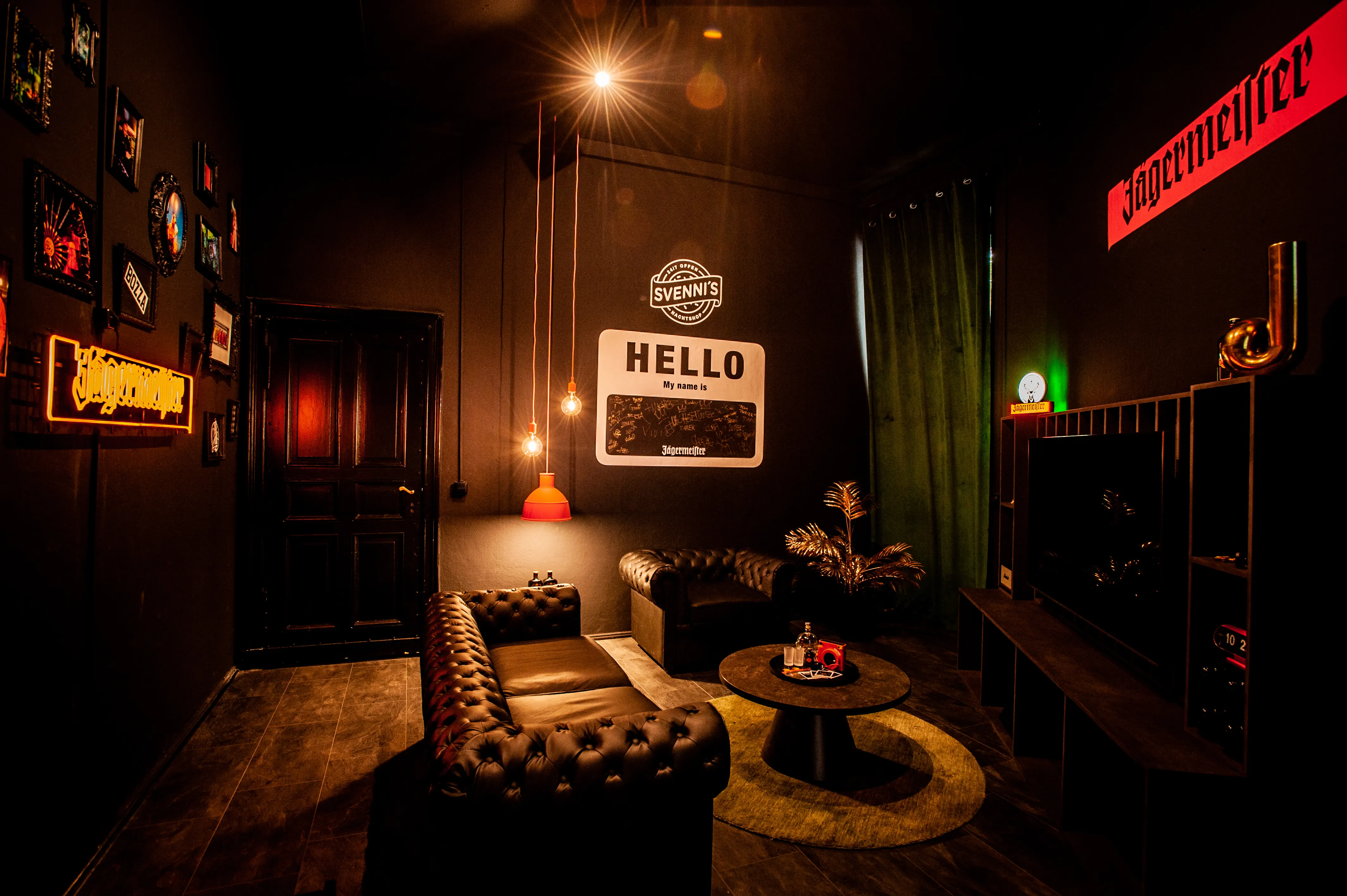 Dark lounge at JÄGERMEISTER x SVENNI'S with brown Chesterfield sofas, a round table, a Hello My Name Is wall graphic, an orange Jägermeister neon sign and framed pictures.