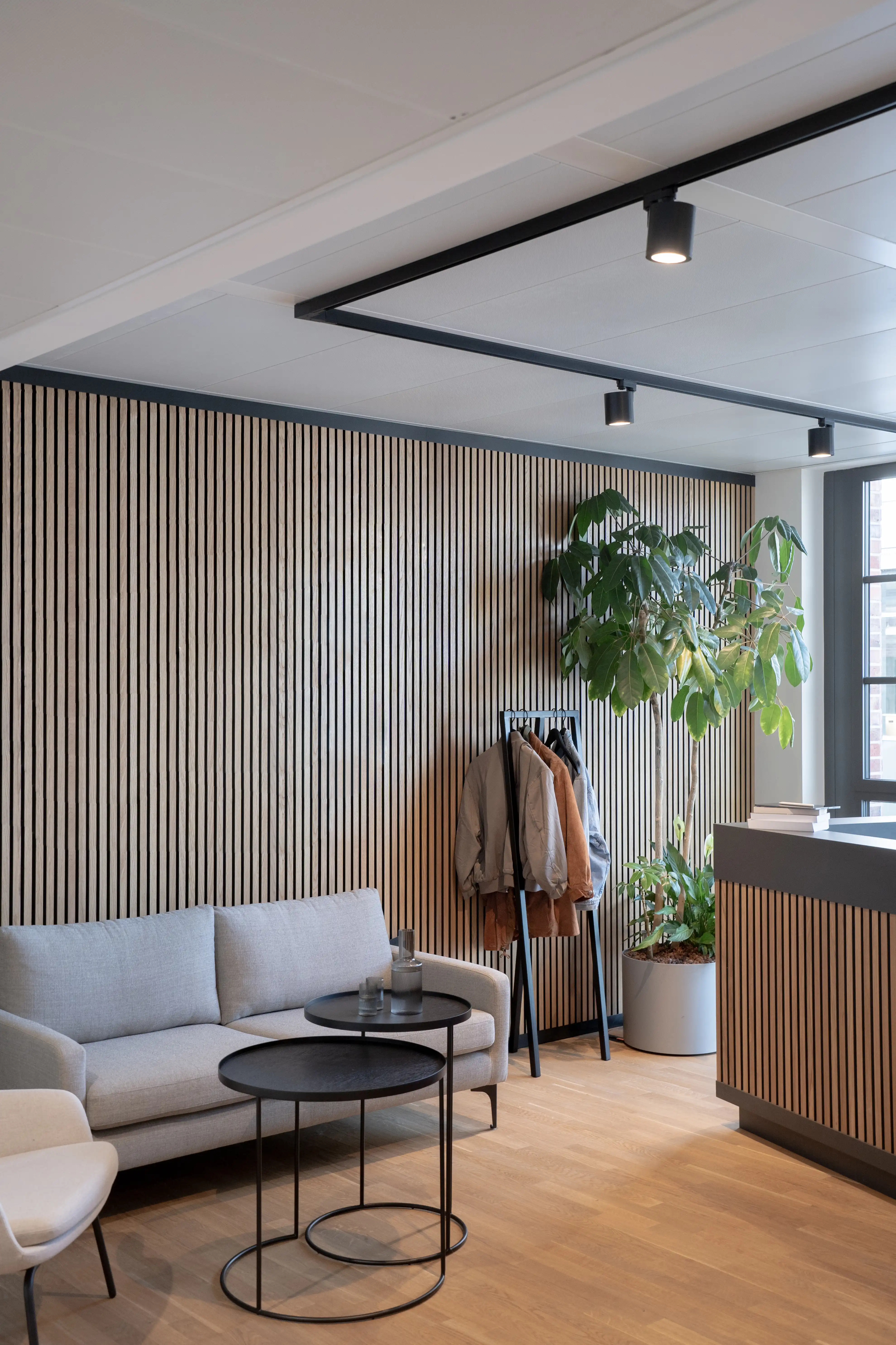 Reception lounge at AI START-UP MUNICH with oak acoustic slat walls, a grey sofa with black nesting coffee tables, a coat rack, a large potted plant and a slat-clad counter.