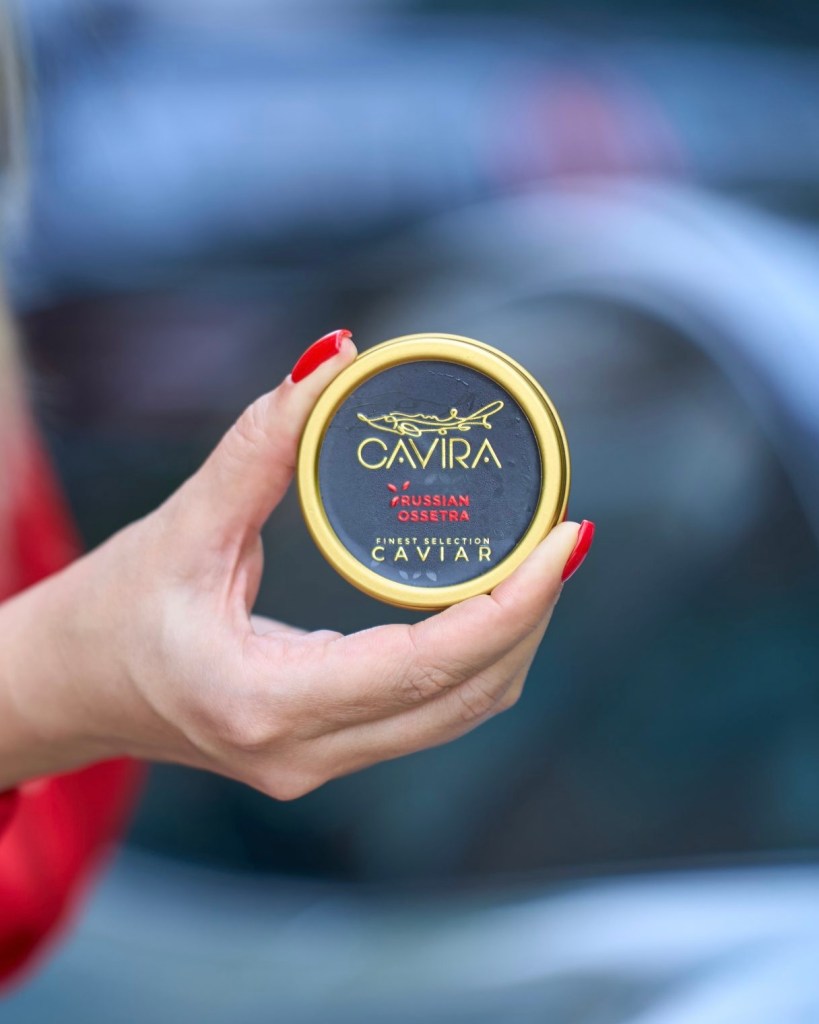 A hand holding a round tin of Cavira caviar, featuring a black label with gold lettering that reads 'CAVIRA', 'RUSSIAN OSSETRA', and 'FINEST SELECTION'.