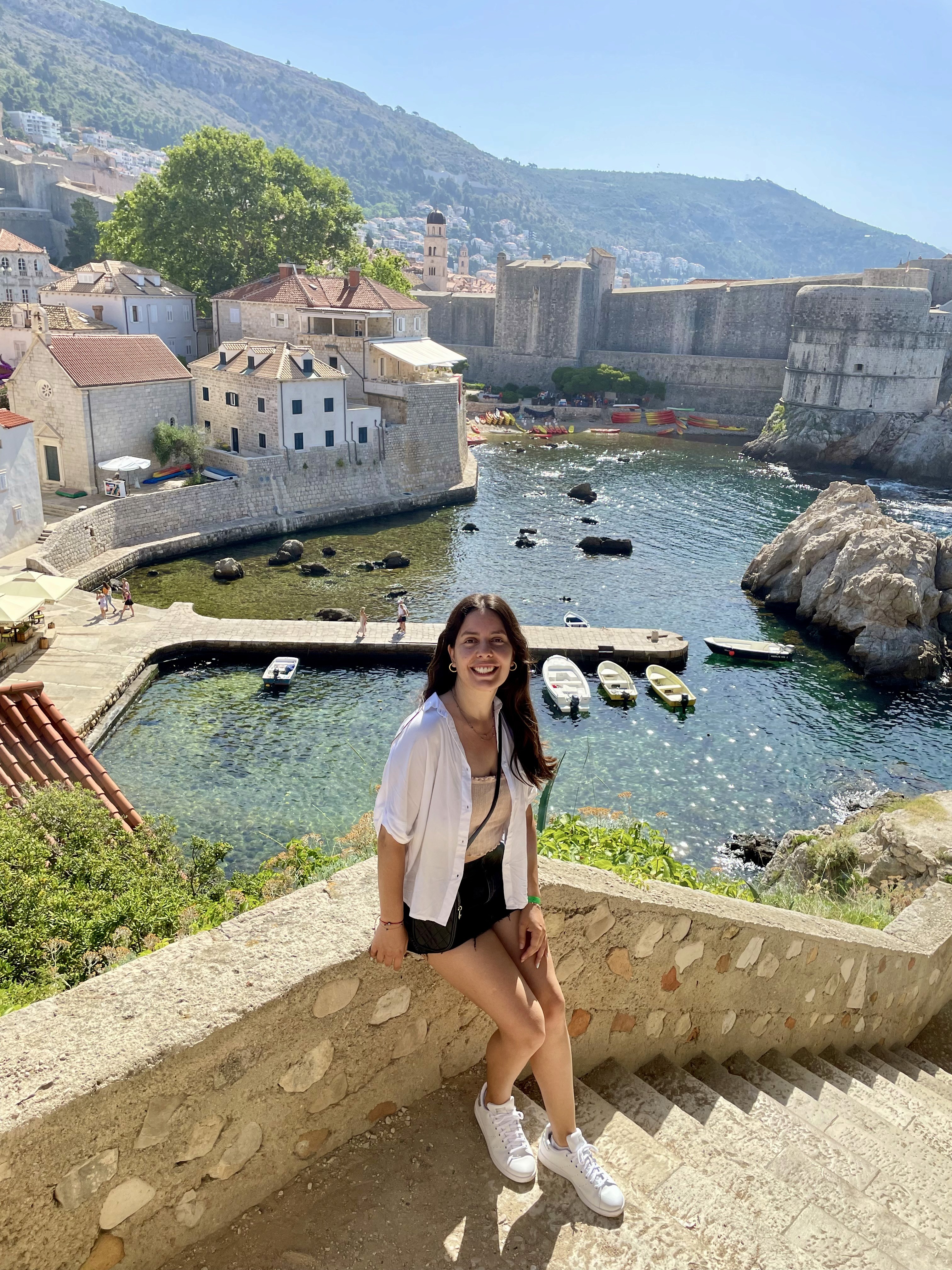 Mayra Coronel sitting on stone steps overlooking a coastal old-town harbor.