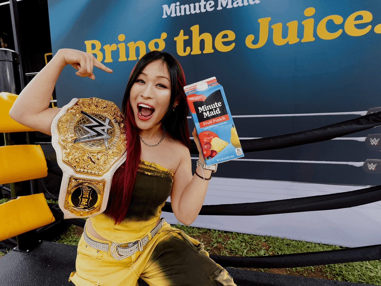 A woman wearing a wrestling championship belt smiles and points to a carton of Minute Maid Fruit Punch while standing in front of a large Bring the Juice sign.