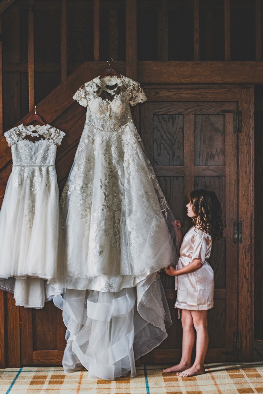 A young girl admires wedding dresses hanging on a wooden door. The dresses are white and intricate, with lace and tulle.