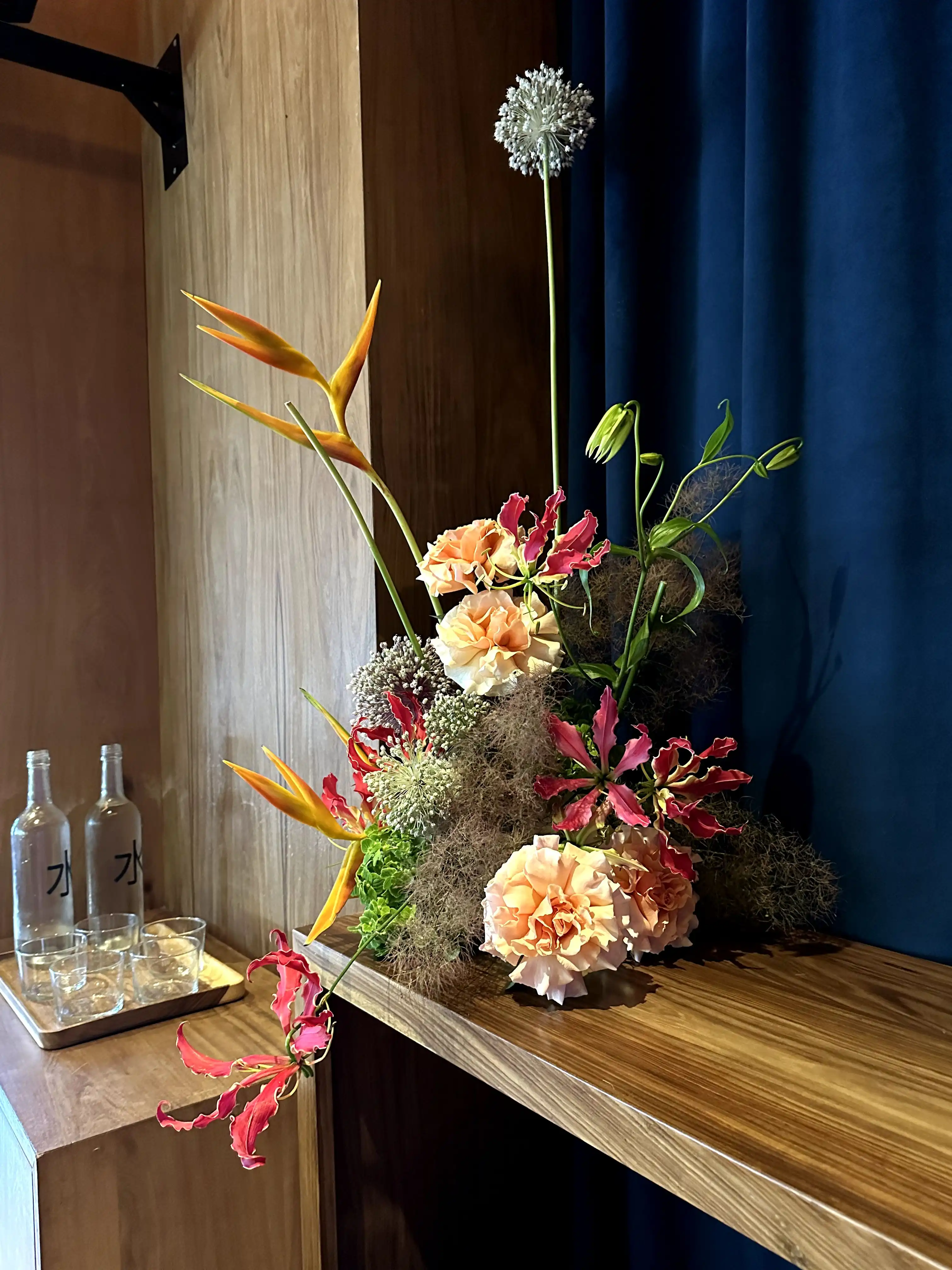 Arreglo floral contemporáneo con aves del paraíso, claveles durazno, gloriosas rojas y flores esféricas lilas sobre una repisa de madera clara frente a una cortina azul, junto a botellas de vidrio y vasos en una bandeja.