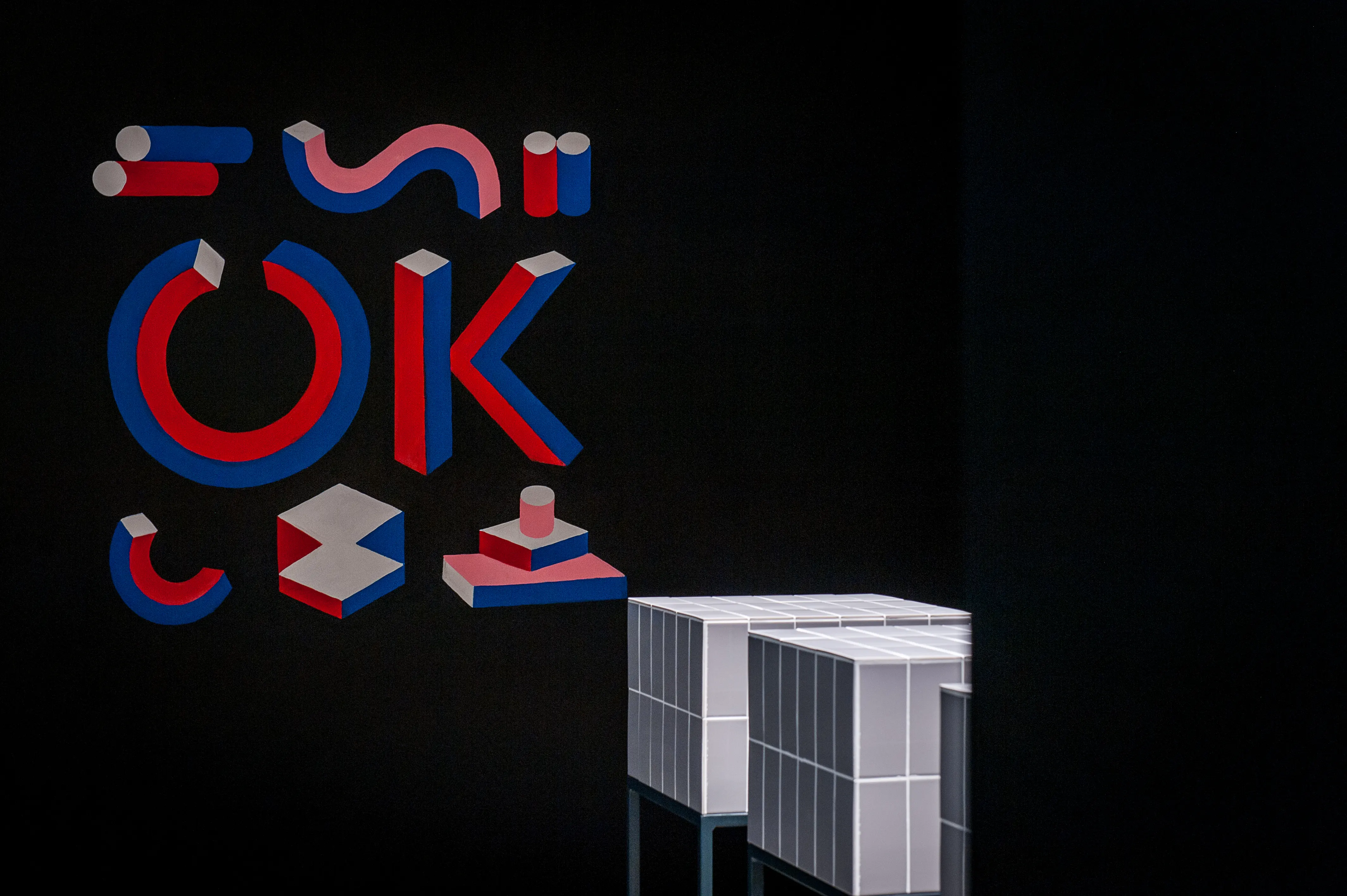 Graphic 3D wall installation spelling OK in red, blue and white geometric shapes on a black wall at the 17 FASHION BRANDS showroom, next to white tiled display blocks.