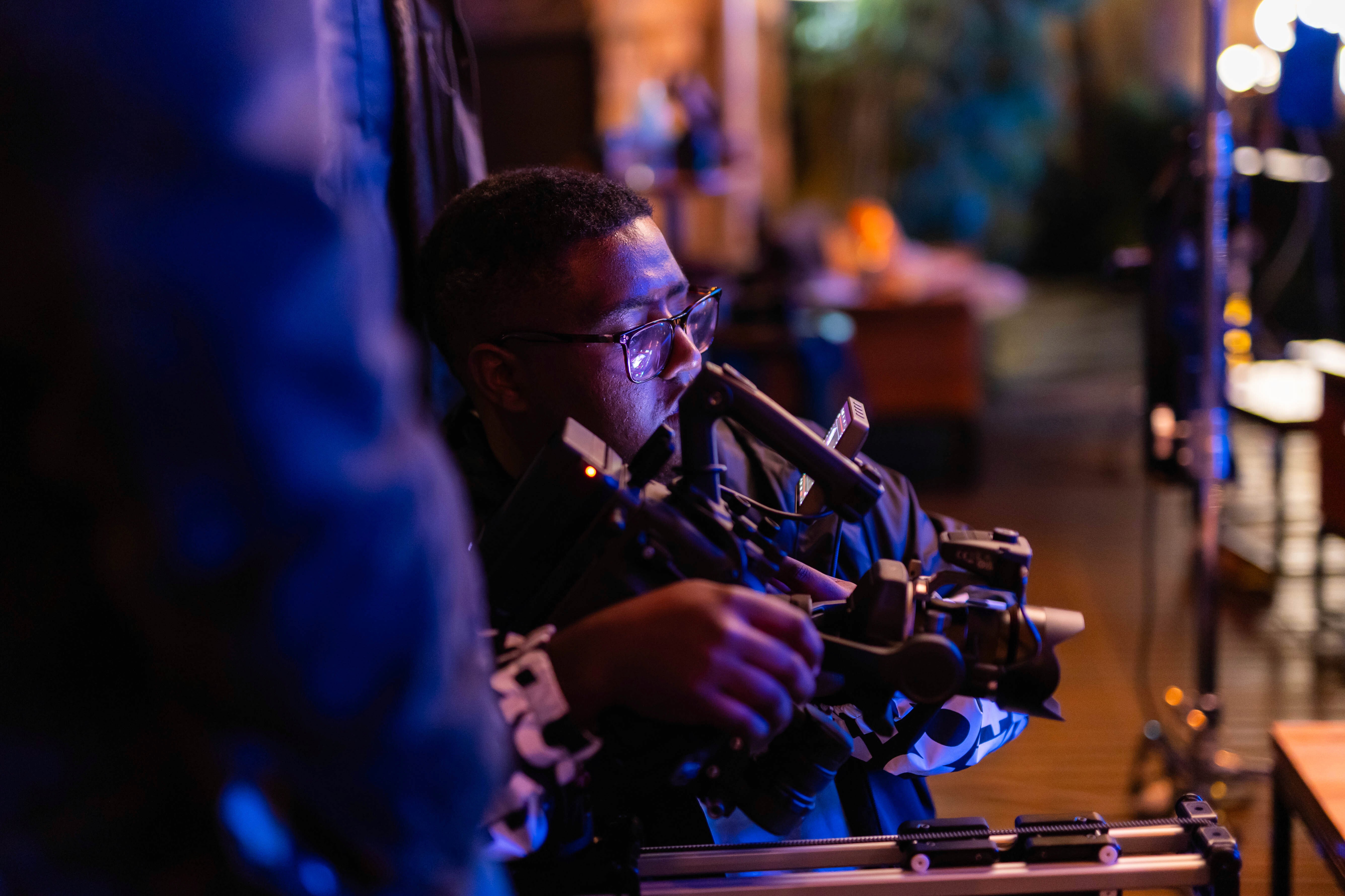 Silhouette of a person with a camera walking towards vibrant orange light under dim blue streetlights on a misty road at night. Mysterious and atmospheric.